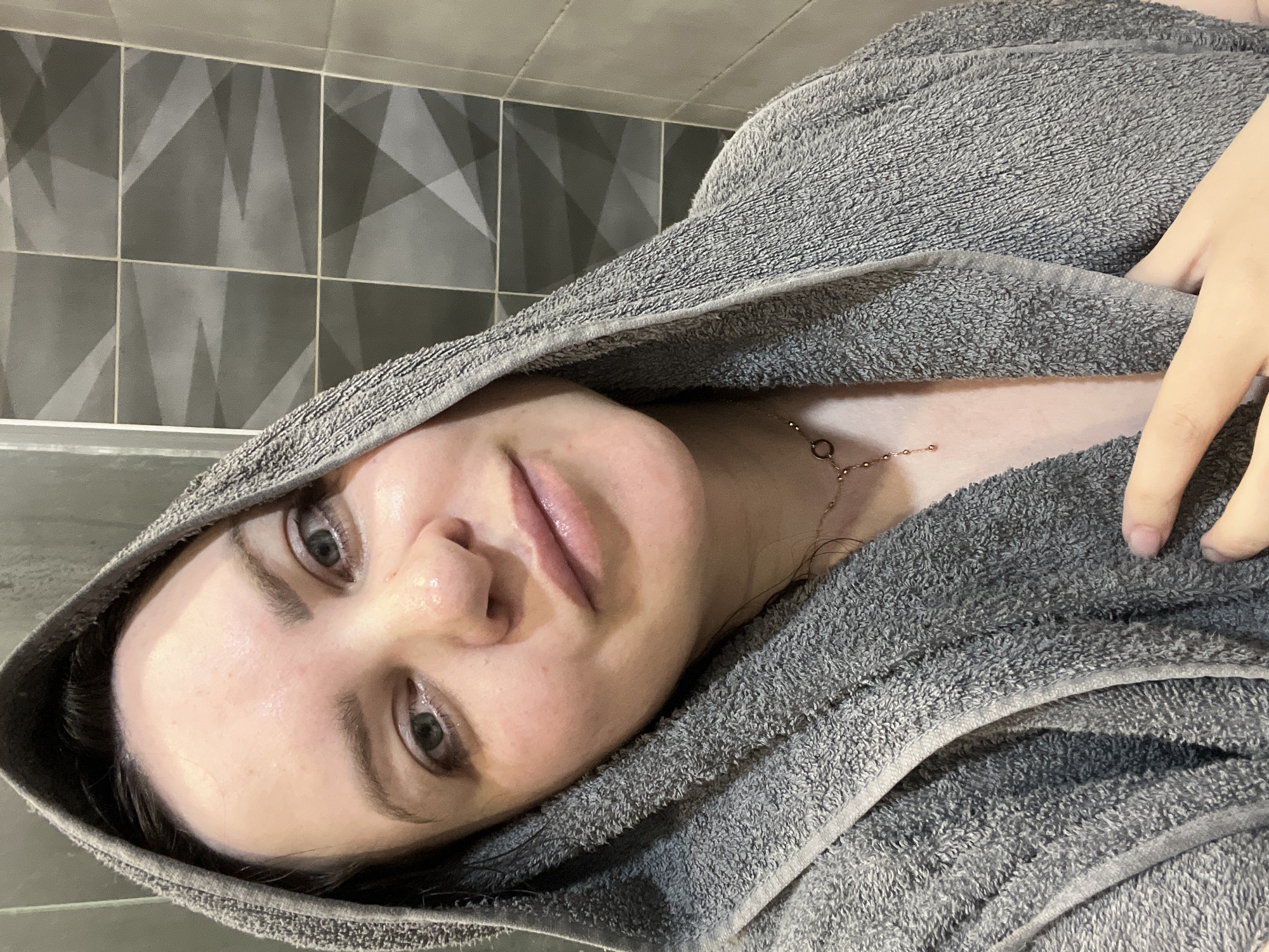 A young adult female with fair skin, wearing a gray towel draped over her head, is indoors. She has a necklace visible on her neck. The background includes gray tilework. It is not possible to determine the location as Noviy, Russia based on the provided image.