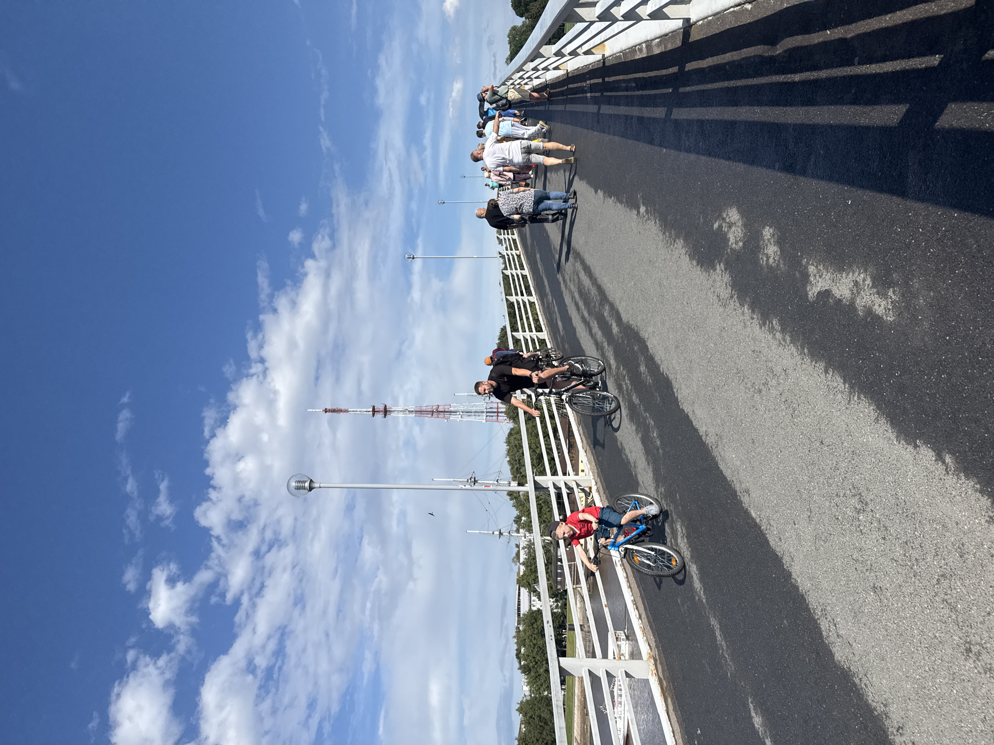 A bright, sunny day with a partially cloudy blue sky is depicted on a bridge in Veliky Novgorod, Russia. A group of people are seen walking and cycling across the bridge. In the foreground, a young boy on a blue bicycle is riding towards the viewer. Behind him, an adult male is cycling on a black bicycle. Further along the bridge, a crowd of people are walking, some in casual attire. In the background, there is a body of water with a white boat, trees, buildings, and a tall radio tower. The bridge has a white railing and a light pole with a glass fixture is visible. The road surface is asphalt with patches of wetness, creating dark and light patches.