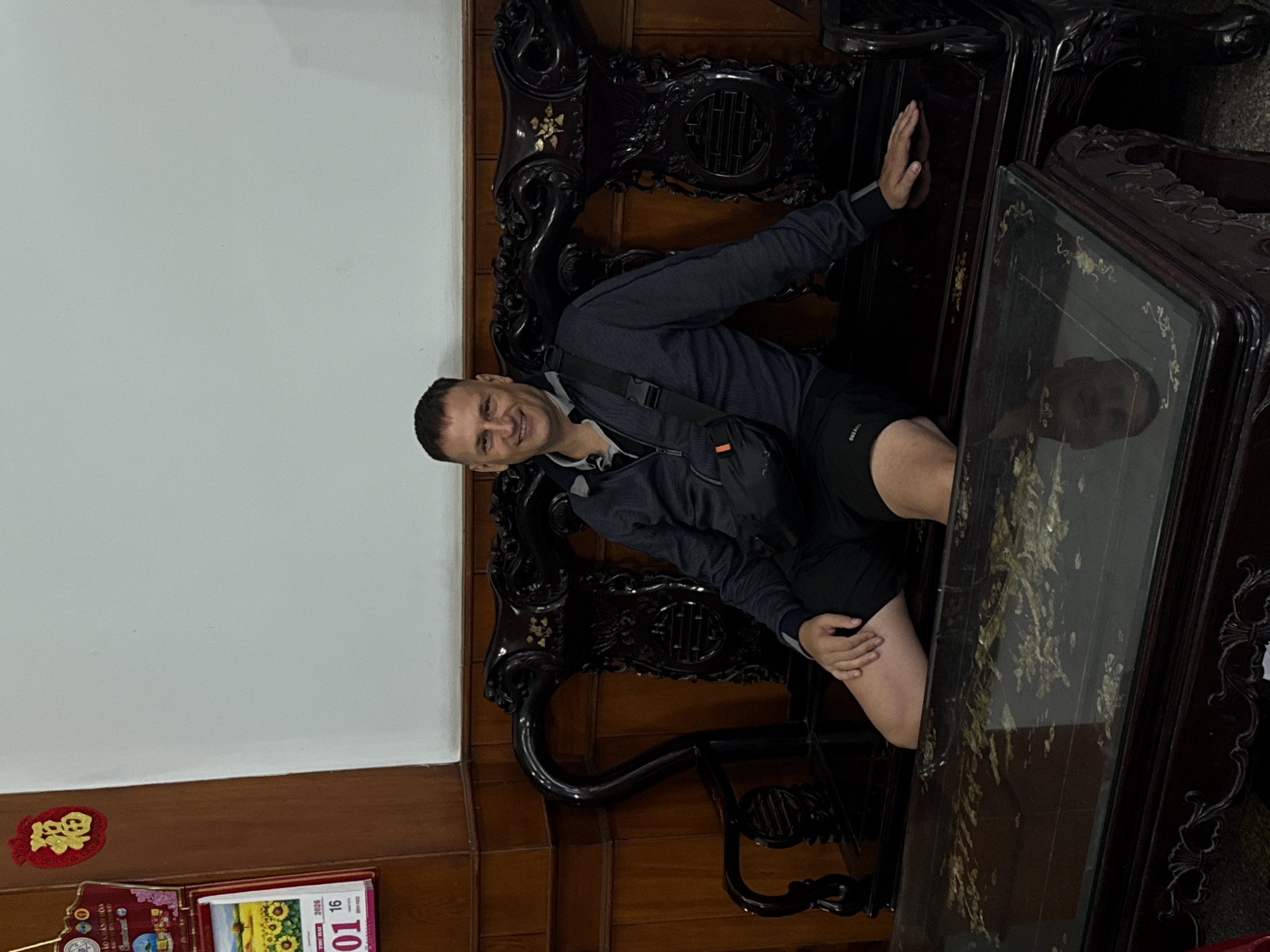 A medium shot shows a smiling adult man sitting on a dark-brown, ornate wooden chair, leaning to his right, while in the city of Tay Nha Trang Ward, Vietnam. The man wears a navy-blue jacket, black shorts, and a black crossbody bag. He is seated beside a dark-brown, ornately-carved wooden table with a reflective, glass top. The table is decorated with gold leaf patterns. In the lower-left corner, there is a red calendar with the date January 16, 2026, visible. The background consists of a white wall above a brown wooden panel. A red and gold decorative item is affixed to the wall above the wooden panel, adjacent to the calendar.