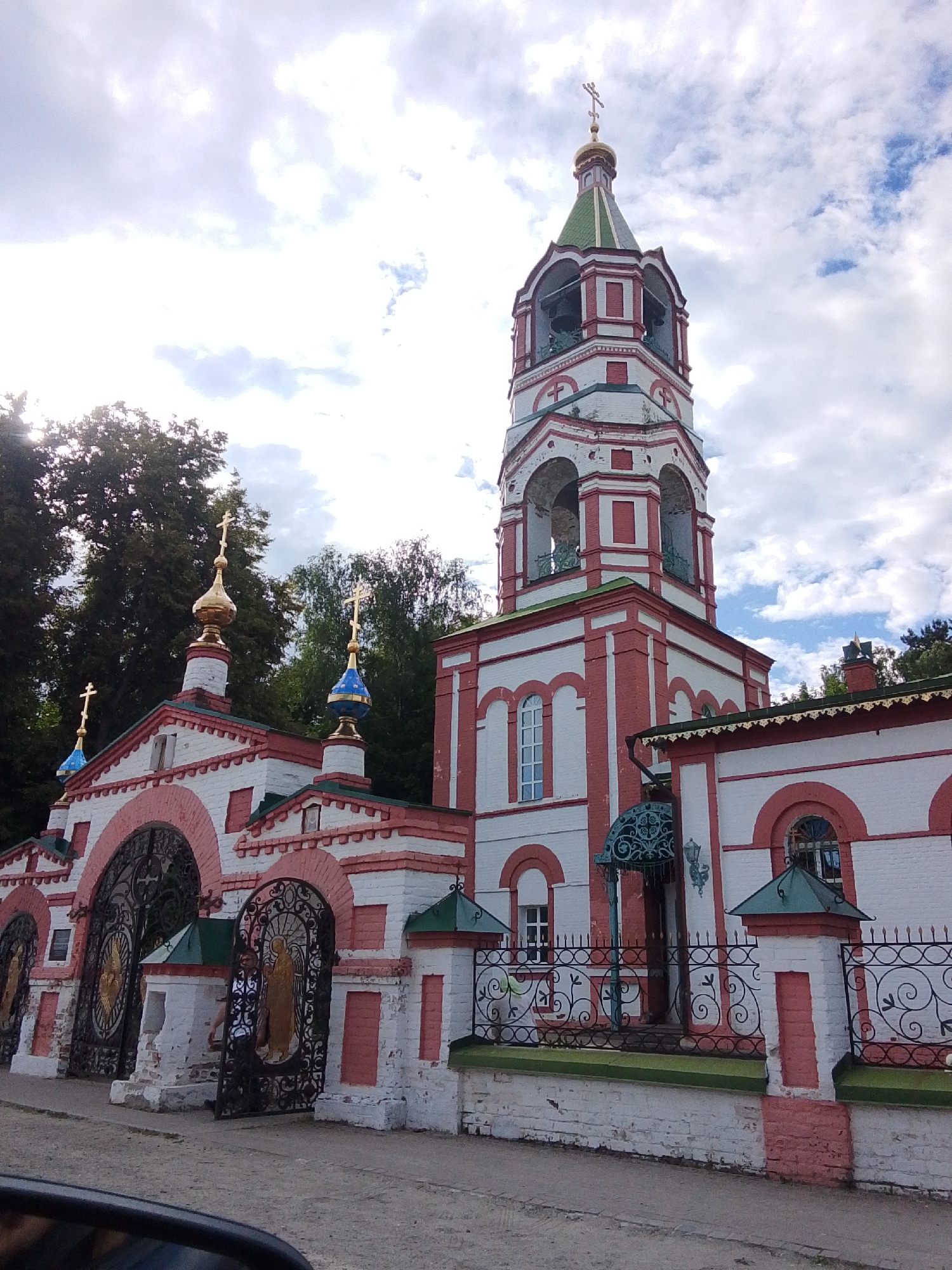 The image shows a frontal view of a Russian Orthodox church and its adjacent bell tower, located in the city of Staryi Pokrov, Russia. The architecture is characterized by white brick walls with red decorative elements and multiple golden domes topped with crosses. The bell tower is tall and octagonal, with arched openings and a green conical roof. In the foreground, there is an ornate wrought-iron gate with decorative elements and golden icons visible behind it. A person is seen standing behind the open gate. The sky is partly cloudy. The environment suggests a sunny day.