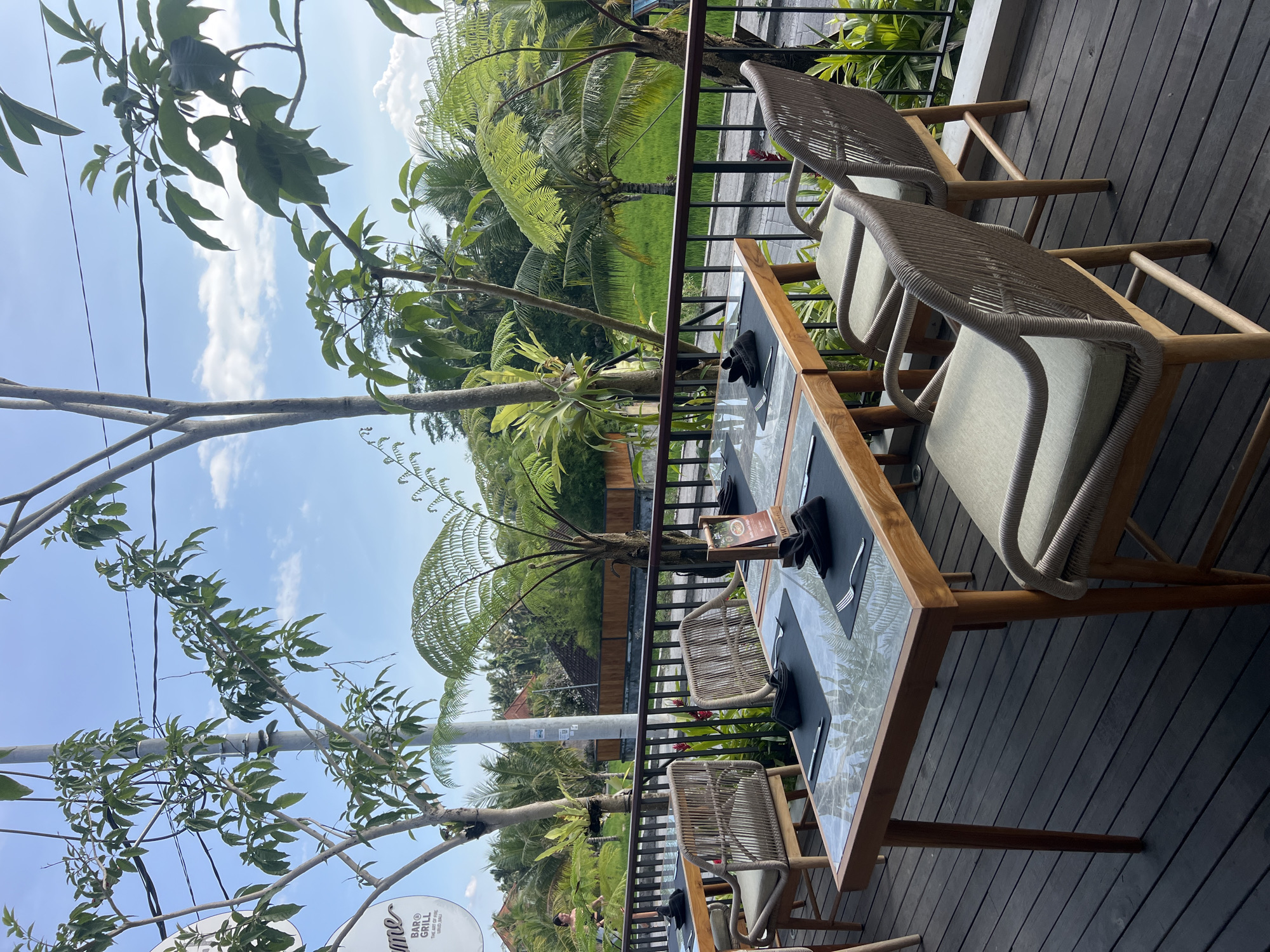 The image shows an outdoor dining area in Tegallalang, Indonesia, with a view of lush greenery and rice paddies. In the foreground, a wooden table with a glass top is set for dining, complete with placemats, cutlery, and folded napkins. Several woven chairs with light-colored cushions surround the table, which is placed on a dark wooden deck. Beyond the railing of the deck, a vibrant green rice field stretches out, bordered by trees and a building. The sky above is blue with scattered white clouds. The overall scene suggests a peaceful and scenic dining experience.