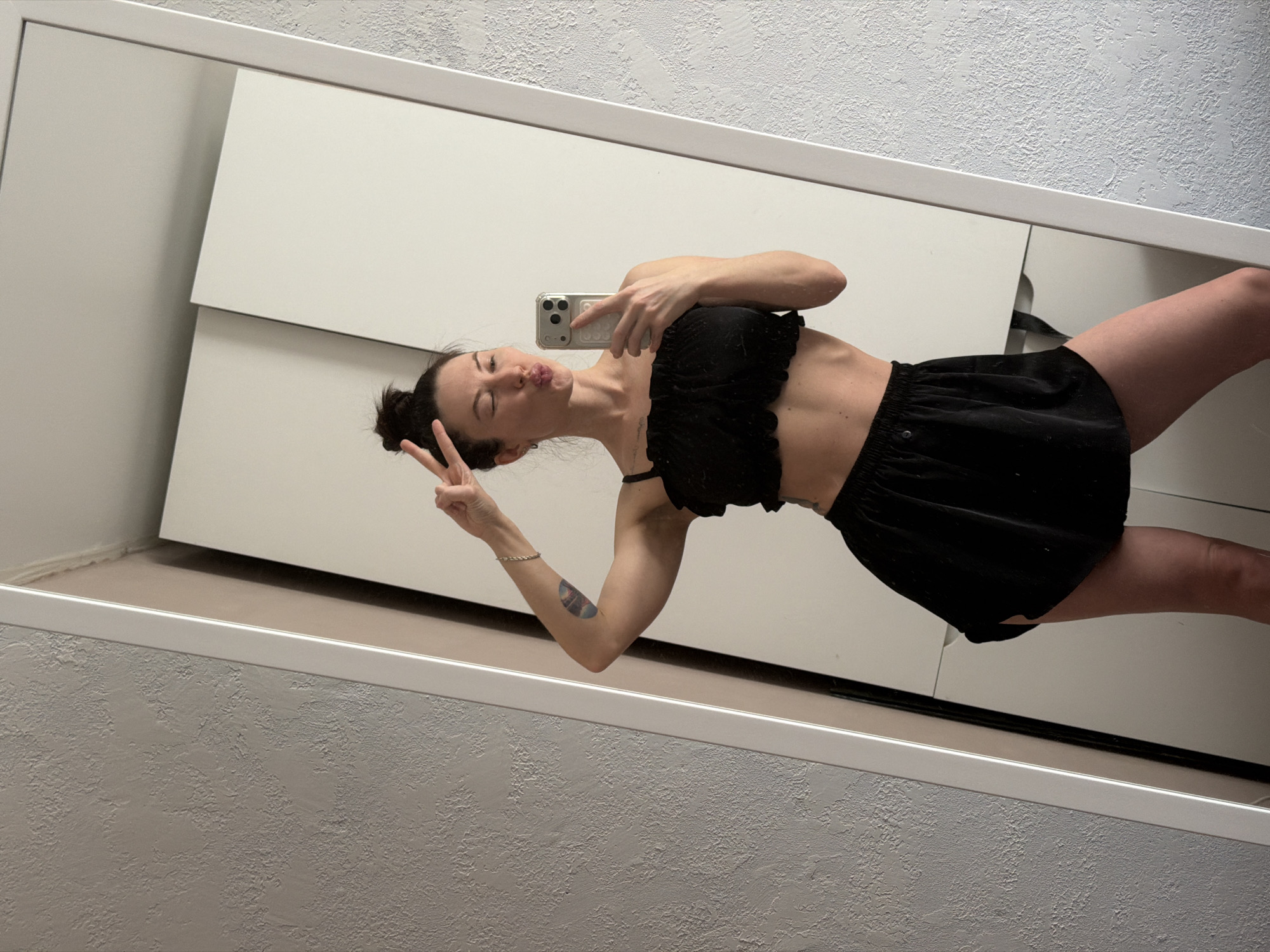 A woman is reflected in a mirror, taking a selfie with a smartphone. She is wearing a black crop top and a black skirt. She is holding up a peace sign with one hand and puckering her lips. The background appears to be a room with white furniture and textured walls. The location is possibly Perm, Russia, though this is not certain from visual information.