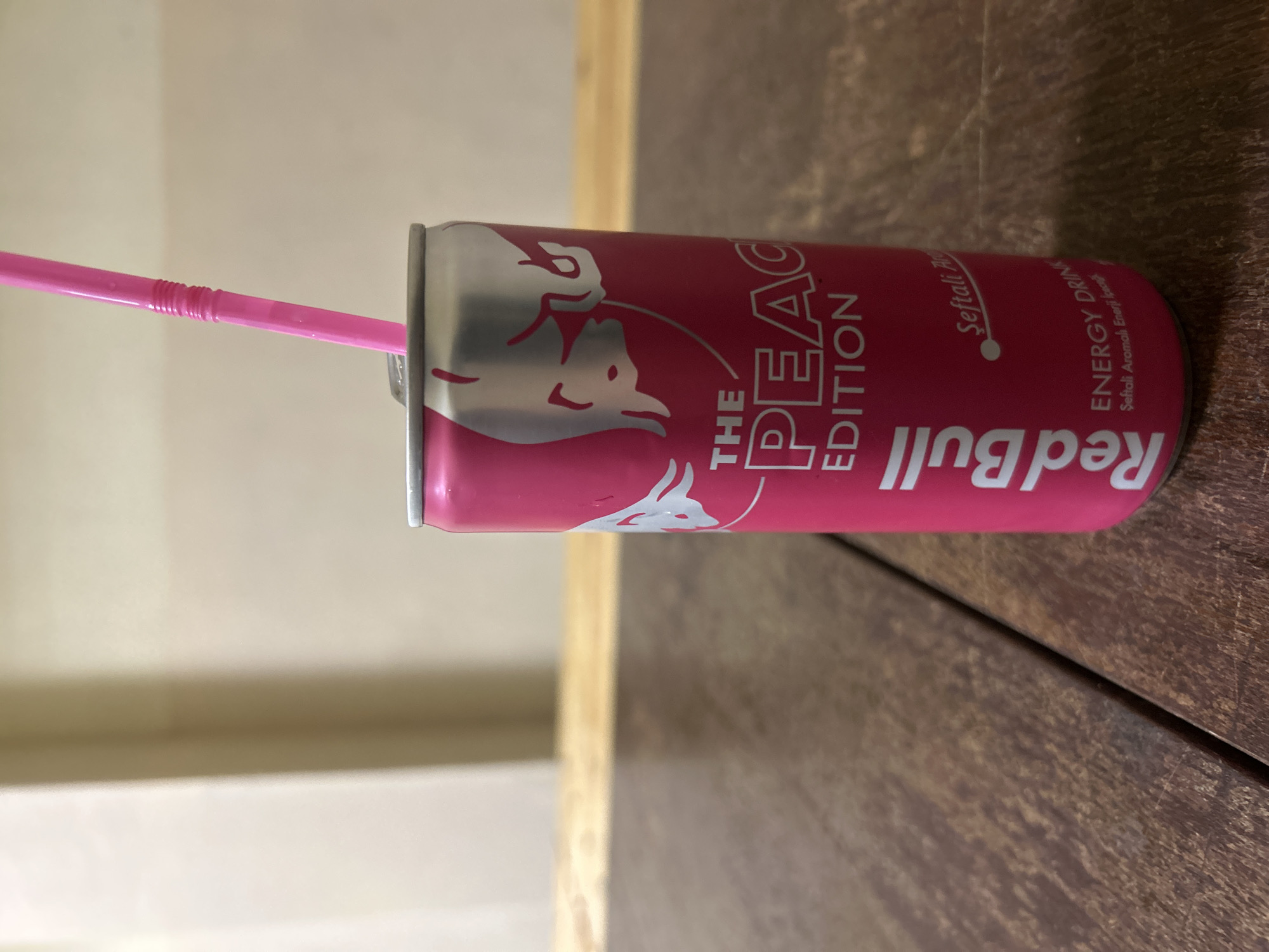 A close-up, slightly angled shot shows a Red Bull "The Peach Edition" energy drink can with a pink straw inserted. The can is primarily pink with silver accents, featuring the Red Bull logo and the text "THE PEACH EDITION." The text on the can also appears to be in Turkish. The can sits on a dark brown, textured wooden surface with a lighter, wooden edge running parallel to it. The background is out of focus, with a gradient of beige to light brown, hinting at a wall or another surface. The location is not indicated in the image.
