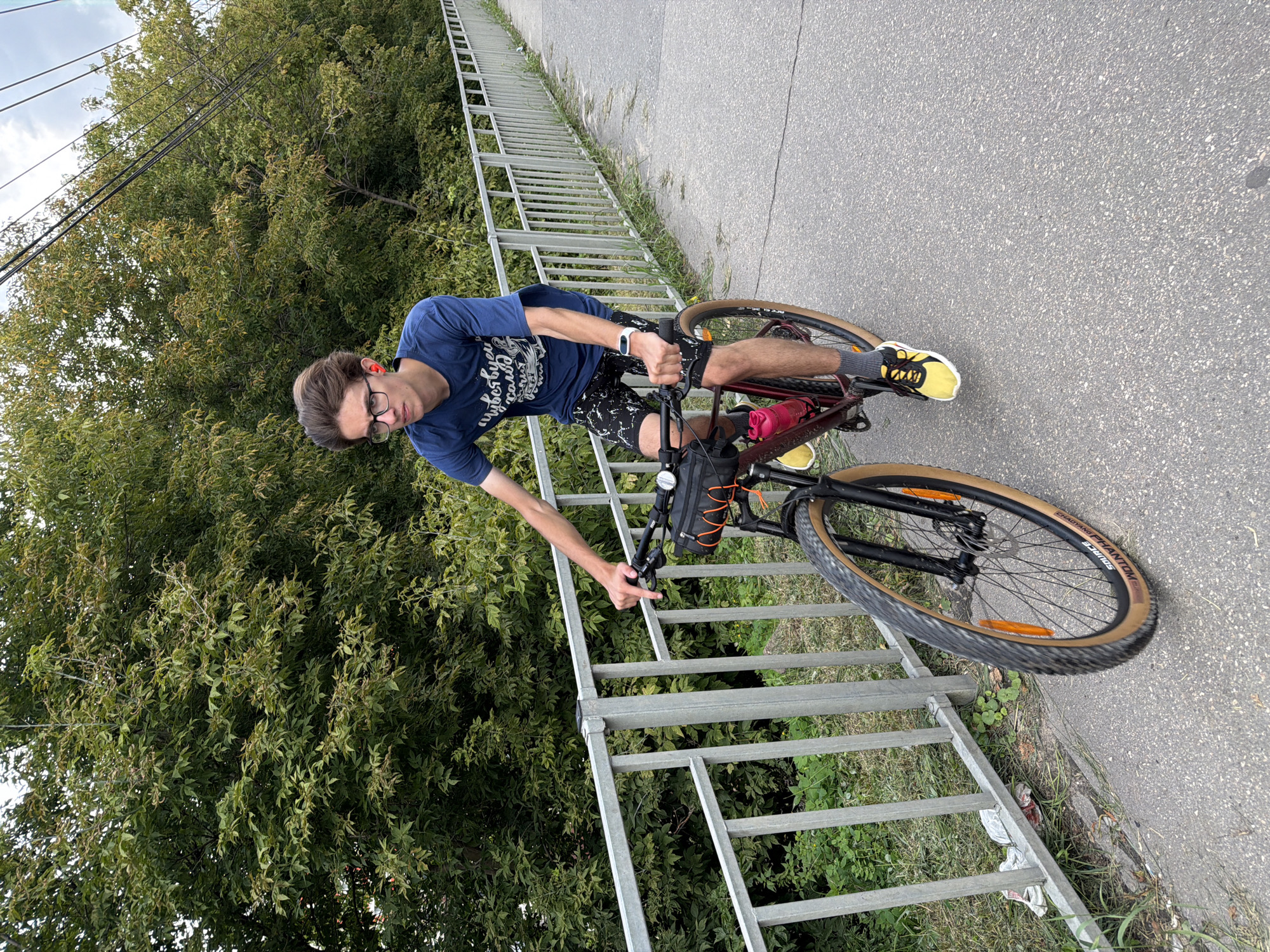 A young man with glasses and red earbuds is riding a bicycle on a paved path in Nizhny Novgorod, Russia. He is wearing a blue t-shirt and black shorts. The path is bordered by a metal fence on the left and lush green trees on the right. The bicycle is dark red with a black bag attached to the handlebars and a pink water bottle on the frame. The man's right hand is on the handlebar, and his feet are on the pedals, wearing yellow and black sneakers. The sky is overcast.