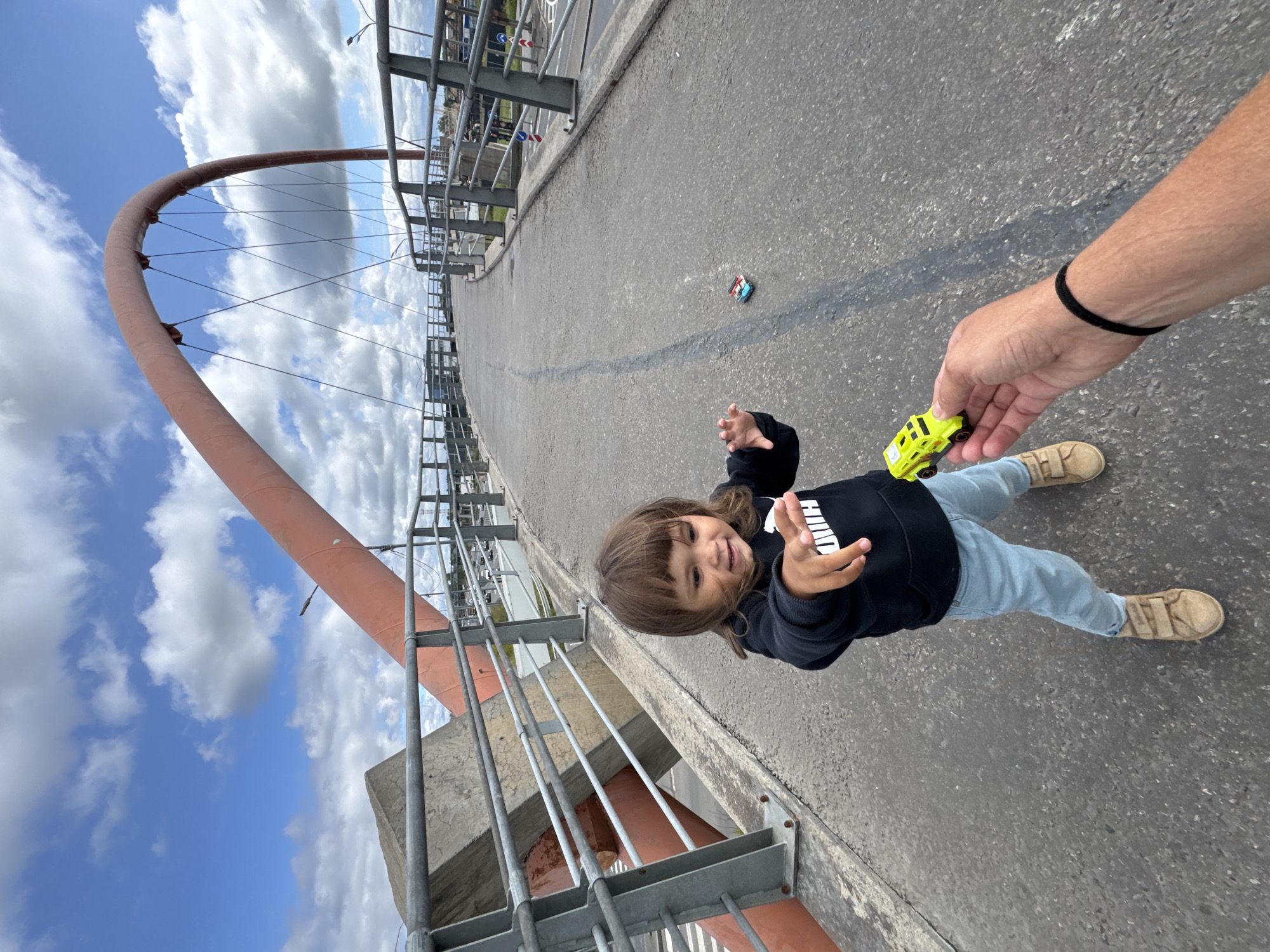 An outdoor, eye-level, full-shot photograph captures a young child on a bridge on a partly cloudy day. The child, a girl with shoulder-length brown hair and bangs, stands on a paved walkway, facing forward with her left arm extended towards the camera. She is wearing a black hooded sweatshirt with a white logo and light blue jeans. Her right hand is outstretched and holding a bright yellow toy truck. A person's arm and hand are visible in the foreground, offering the toy. In the background, a red arched bridge structure spans the walkway, supported by cables. The walkway curves to the right, with metal railings on either side. In the distance, through the railings, a road with cars and buildings can be seen. The scene appears to be the bridge leading to the Gazprom Arena in St. Petersburg, Russia.