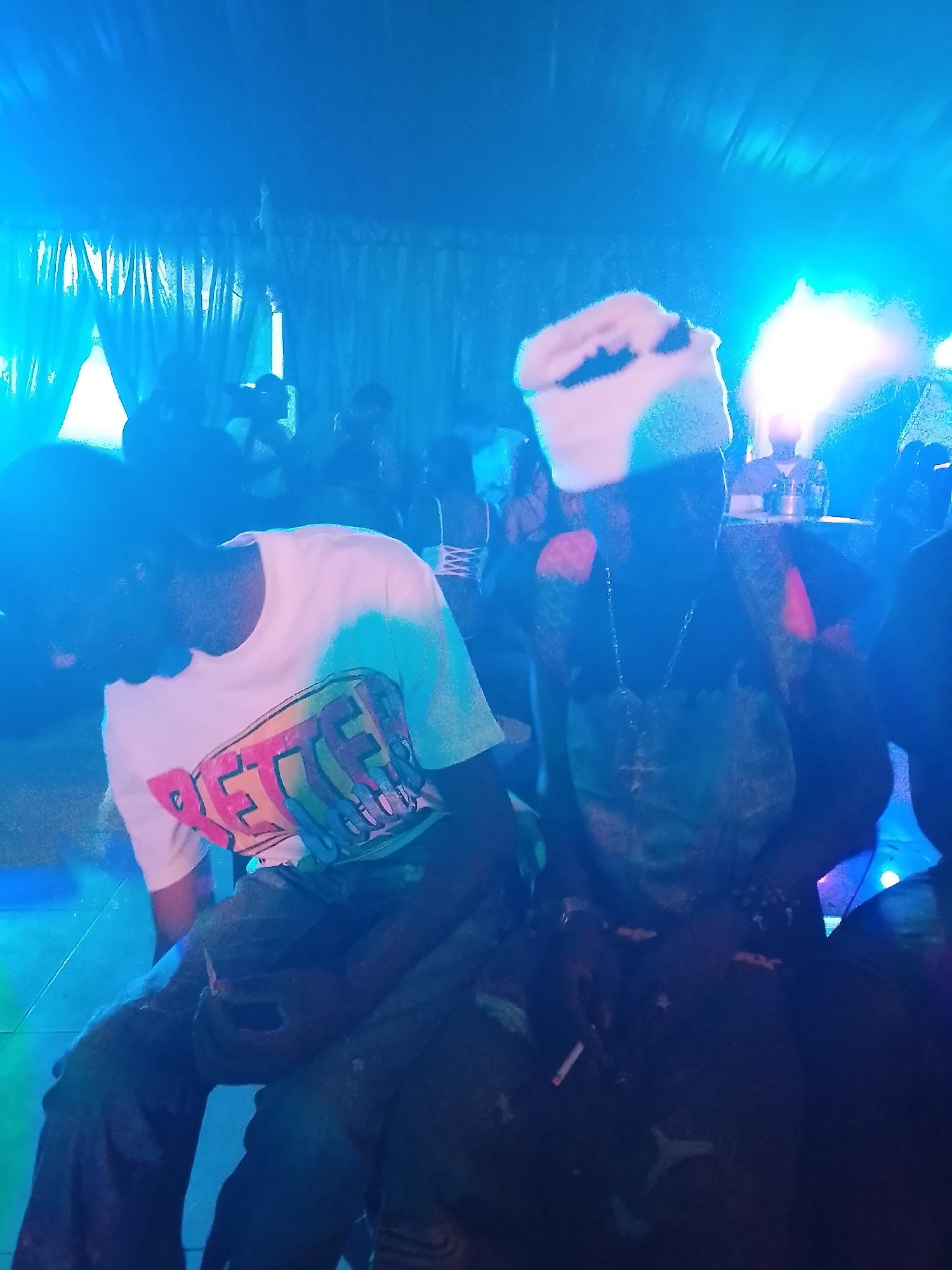 A nighttime indoor event is depicted. Two people are featured prominently, with others in the background. The individual on the left wears a white t-shirt with a large, colorful graphic design. The person on the right sports a hat and a dark outfit, with a necklace visible around their neck. Other individuals are partially visible in the backdrop. The environment is dimly lit, with blue lighting casting shadows and creating a vibrant yet somewhat obscured atmosphere.