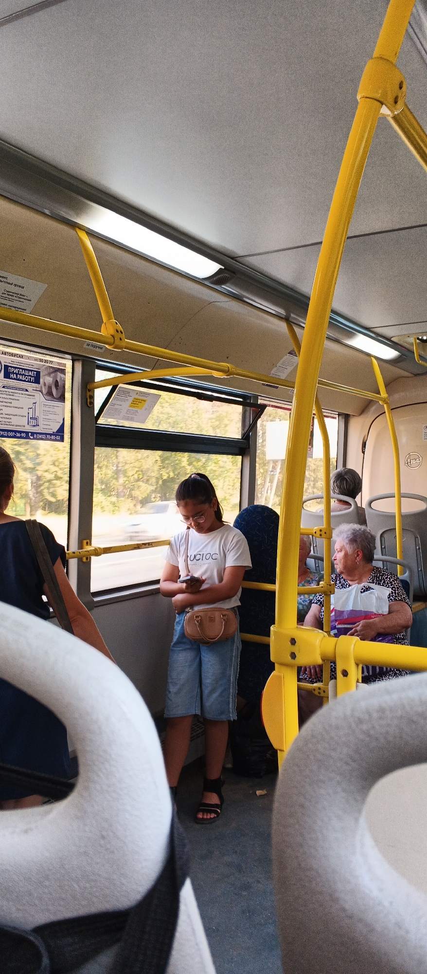Inside a public bus, likely in Izhevsk, Russia, a young woman with dark hair pulled back in a ponytail stands near a window, looking down at her smartphone. She is wearing a white t-shirt with a graphic, denim shorts, and a small brown crossbody bag. To her left, a woman with dark hair is partially visible, facing away from the camera. To her right, an elderly woman with grey hair sits facing forward, wearing a patterned top and holding a shopping bag. The bus interior features yellow handrails, fluorescent lighting, and windows showing trees and a passing vehicle outside. The floor is dark grey.