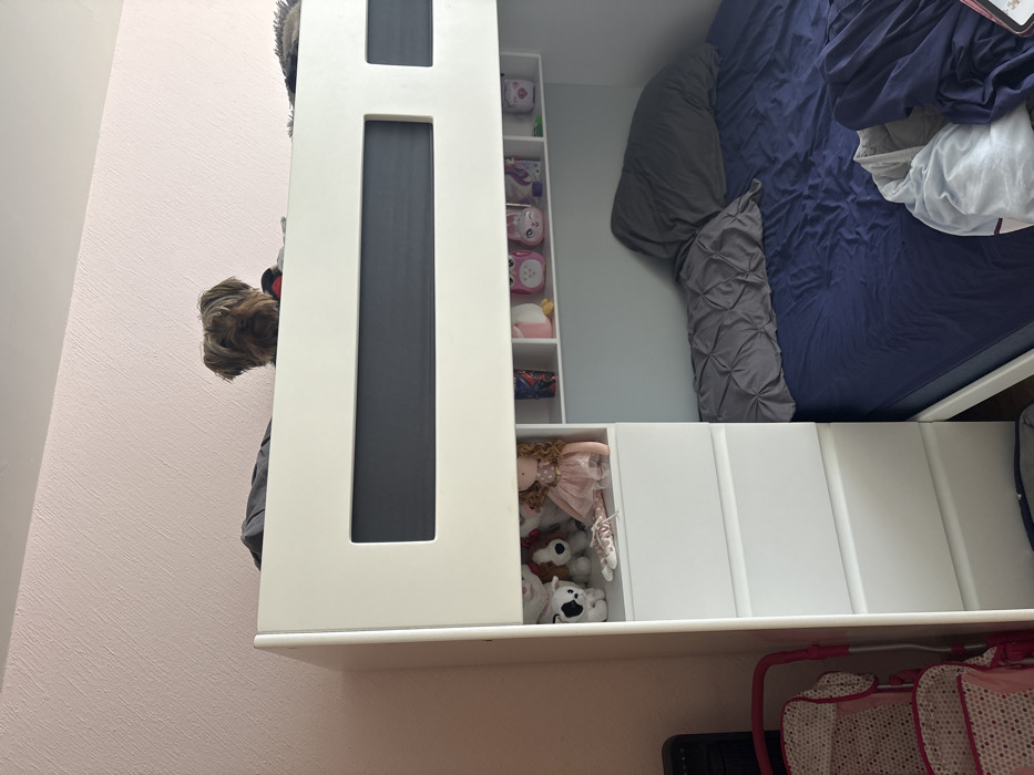 The image displays an interior room, likely a bedroom, focused on a white bunk bed. A Yorkshire Terrier dog is visible atop the upper bunk, with its head and upper torso peering over the edge of the bed frame, facing forward. The upper bunk's frame incorporates a dark grey horizontal panel. The lower bunk is made with a navy blue fitted sheet, a rumpled grey duvet, and two grey pillows. Integrated into the bunk bed structure on the left side is a white dresser with multiple drawers and an open shelving unit above the lower bunk. This shelf displays a light pink doll, several plush toys (including a black and white dog and a brown dog), and various small items such as cartoon-themed cups, a pink owl figurine, a white pig figurine, a white unicorn figurine, and other small white and green objects. To the far left, a portion of a black, vertical appliance is visible. In the lower left foreground, part of a pink and white patterned play stroller is present. The room's background wall is a textured light pink. The specific location within León, Mexico, cannot be determined from the visual information provided.