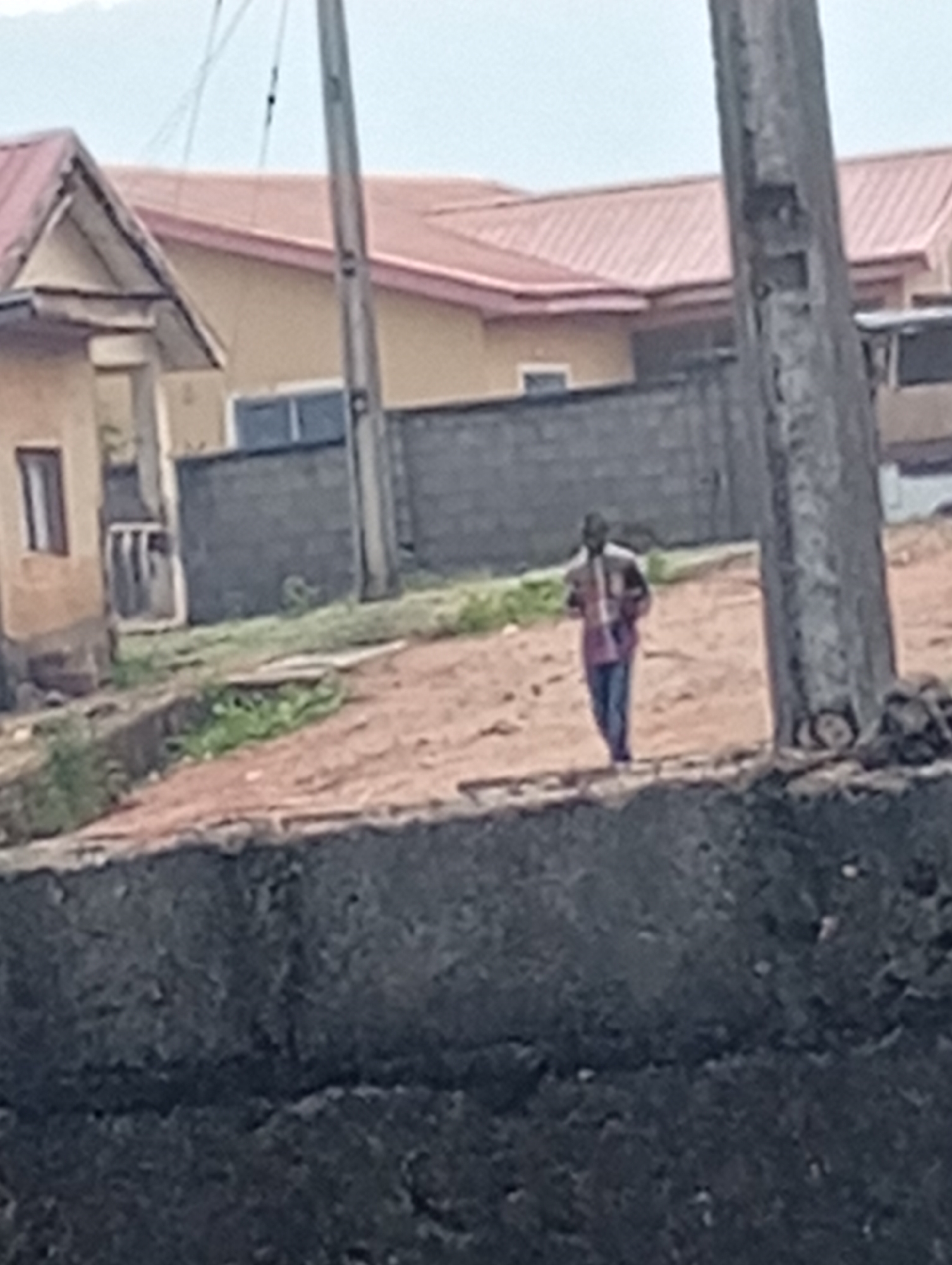 The image depicts an outdoor scene in Adankolo, Nigeria, featuring a lone individual walking along a dirt path under an overcast sky.
In the mid-ground, an adult male figure is seen walking towards the left. He is dressed in a patterned, long-sleeved shirt, possibly plaid or checked, and dark trousers. The ground is predominantly reddish-brown earth, with scattered patches of green vegetation, particularly to the left of the path.
The background features several buildings with distinct red or orange corrugated metal roofs and light-colored walls, characteristic of residential architecture in the region. A prominent house on the far left has a gabled roof. A dark, low concrete block wall runs horizontally across the mid-background, separating the path from the more distant structures. On the right side of the frame, a tall, slender utility pole extends from the bottom of the image towards the top, with faint wires visible against the sky.
The immediate foreground shows a rough, dark concrete structure, possibly a low wall or embankment. The sky is uniformly light grey, indicating an overcast or cloudy day. There is no specific activity beyond the person walking, and no visible text in the image. The scene conveys the impression of a typical street or pathway in a residential area.