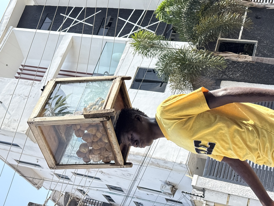 A male individual is observed in Igboefon, Nigeria, walking outdoors under bright daylight, balancing a rectangular display case on his head. The case is constructed from light-colored wood framing and glass panes, containing numerous round, light-colored food items, likely baked goods, on the left side, and smaller, irregular yellowish items, possibly fried snacks, on the right. The individual wears a bright yellow short-sleeved t-shirt with a dark, stylized logo on the chest. His head is slightly tilted backward, and his eyes are closed or squinted, indicating concentration on maintaining balance.
The background consists of two multi-story buildings. The structure to the left is predominantly white with multiple windows and an externally mounted air conditioning unit, partially obscured by a grey gate. The building to the right is a more modern design, featuring white and dark grey/black sections, accented with contrasting white vertical and diagonal line patterns. This building includes dark-tinted windows and a balcony with a glass railing. Several green palm trees are positioned in the mid-ground. Overhead, utility lines traverse the frame against a clear, light blue sky.