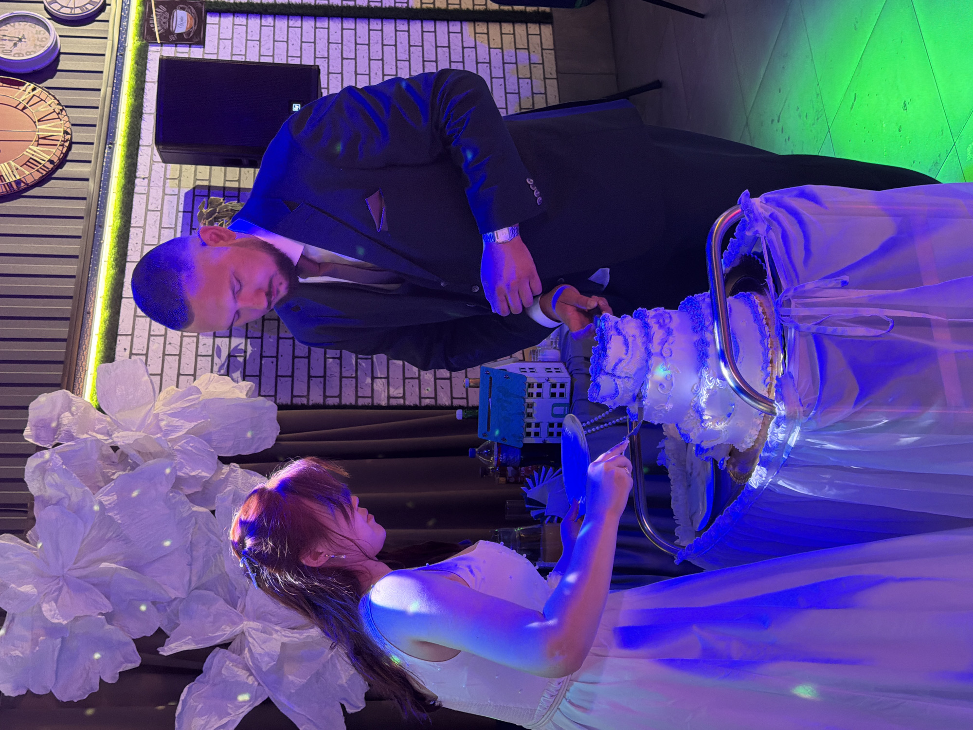 In Chernushka, Russia, at what appears to be a wedding reception, a woman in a white dress and a man in a dark suit are cutting a multi-tiered white cake. The woman is on the left, holding a knife and a plate, with her hair pulled back and adorned with a hair clip. The man is to her right, also holding a knife. The cake is decorated with white frosting and blue accents. They are standing behind a table draped with a white tablecloth. In the background, there are large white floral decorations, a brick wall, and some clocks on the wall. The lighting is a mix of blue and green, suggesting a festive atmosphere.