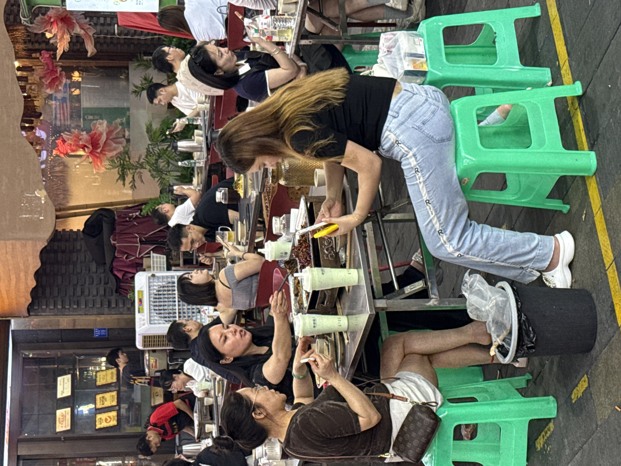 The image depicts a lively evening scene at an outdoor or semi-outdoor dining establishment, likely a street food market or restaurant alley in Liangjiang, China. The area is bustling with numerous people, primarily young adults, gathered around long, narrow tables.
The subjects include a multitude of diners, tables, and bright green plastic stools. In the foreground, a young woman with long, light brown hair, dressed in a black top and light blue jeans with a repeating 'R' pattern on the side, stands leaning over a table, holding yellow tongs. She appears to be serving or preparing food. Other individuals are seated, engaged in various activities such as conversing, eating, and looking at their mobile phones.
Notable details include the time of day, which is evening or night, indicated by the artificial lighting and overall ambiance. The setting suggests a casual, communal dining experience. The tables are filled with small dishes, food containers, and plastic cups holding a light green beverage. A white cooler or fan unit is placed between tables. In the lower right, a person's legs are visible next to a black cylindrical bin with a plastic bag liner, and a small brown handbag with a monogram pattern rests on a green stool. The background features decorative, illuminated red and pink floral structures and blurred storefronts with visible signs displaying Chinese characters. The overall atmosphere is vibrant and social.