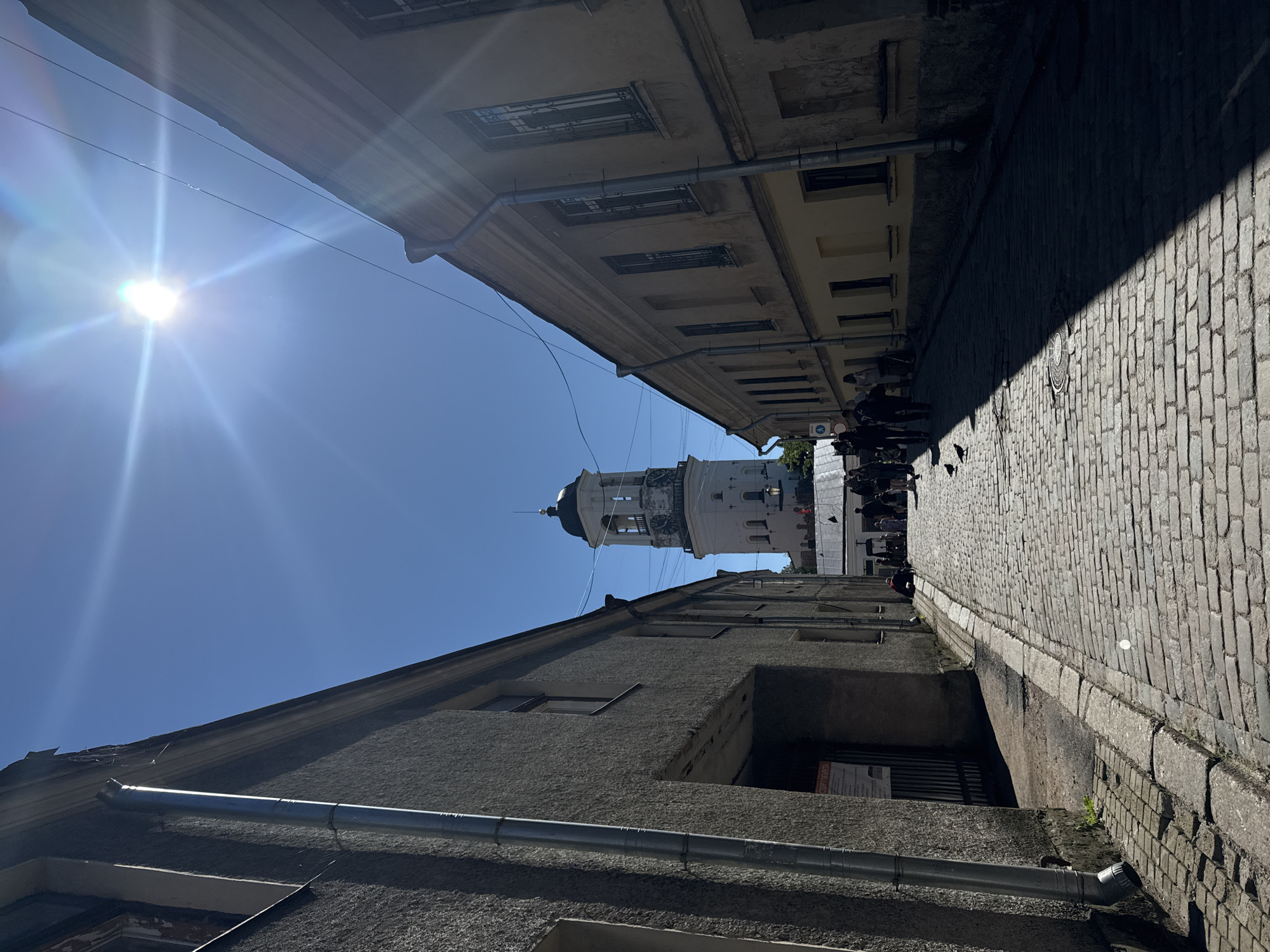 A street view shows a narrow cobblestone street lined with buildings on both sides, leading towards a clock tower in the distance under a clear blue sky. The sun is high and bright, casting a strong shadow across the street from the building on the right. Several people are walking up the street, some in groups, and a few pigeons are on the cobblestones. The building on the left is gray and appears to be older, with a metal downspout running vertically along its facade. The building on the right is a lighter color and has several tall windows with barred exteriors. The clock tower is white with a dark steeple and a clock face visible. The scene is framed by the architecture, emphasizing the path leading towards the prominent tower.