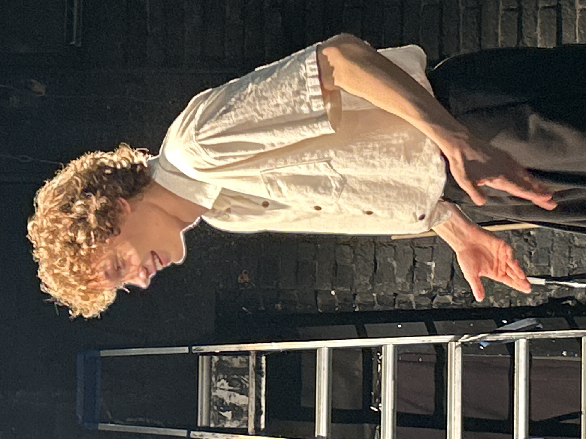 The image shows a person with curly blonde hair and fair skin, facing to the left and looking down. They are wearing a light-colored, short-sleeved button-up shirt with two pockets. Their right arm is bent, and their hand is open, palm facing downwards. The background consists of a dark, rough-textured surface, possibly brick or stone, with some lighter-colored elements and reflections. In the lower portion of the image, a metal ladder with blue accents is visible. The lighting appears to be artificial and casts shadows, suggesting an indoor or nighttime setting. The overall mood is somewhat dramatic due to the lighting and pose. No text is visible in the image.