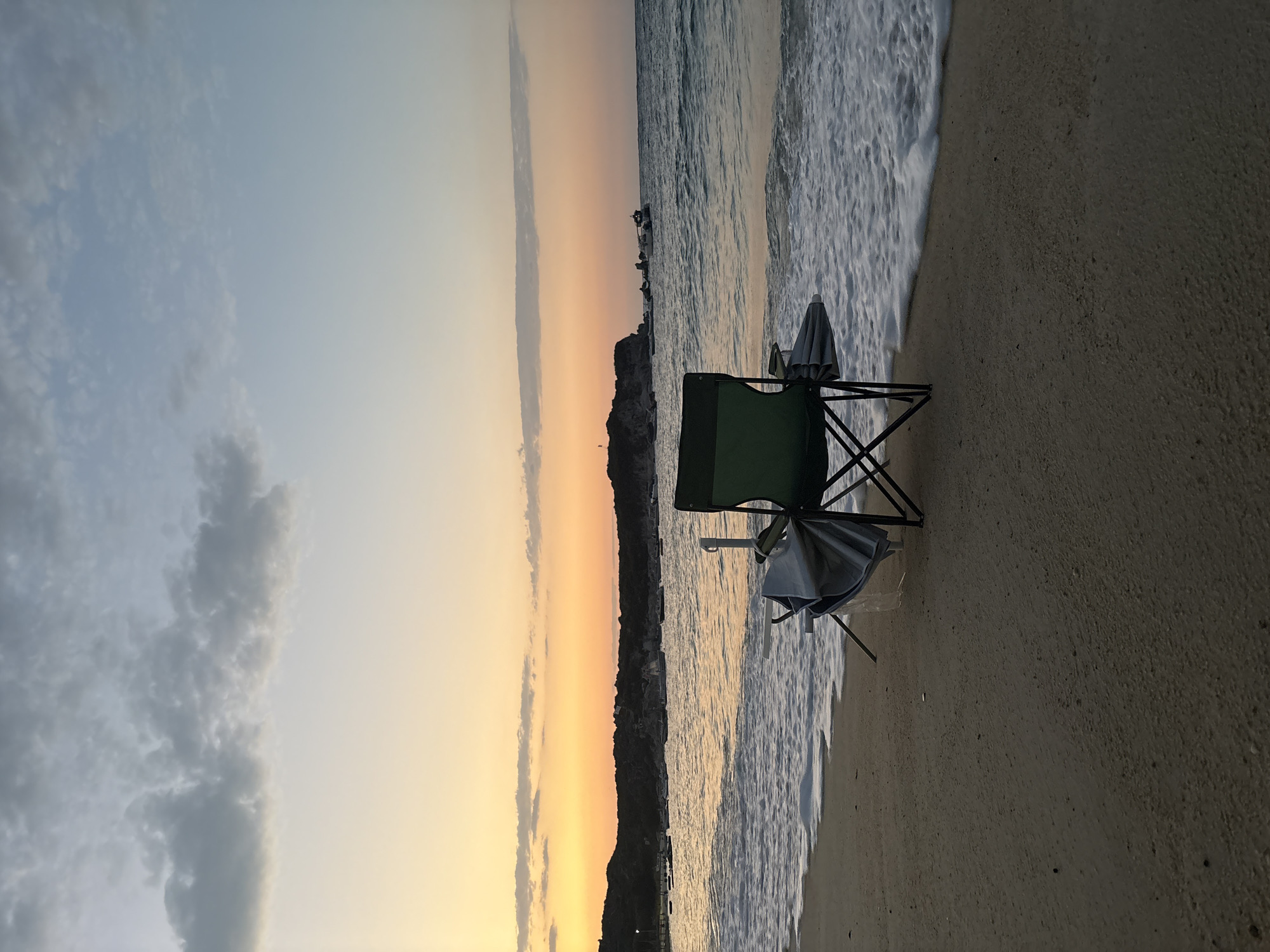 An outdoor scene captures a beach at sunset. In the foreground, a green folding chair sits on the sandy shore, with a partially collapsed umbrella leaning against its side. The ocean laps at the sand with gentle waves, creating a white foam line. In the background, a rocky coastline is visible, with trees and some structures dotting the landscape. Several boats are anchored in the water. The sky displays a gradient of colors from warm orange and yellow at the horizon to soft blue and gray clouds higher up. The location is the city of Toroni Municipal Unit, Greece.