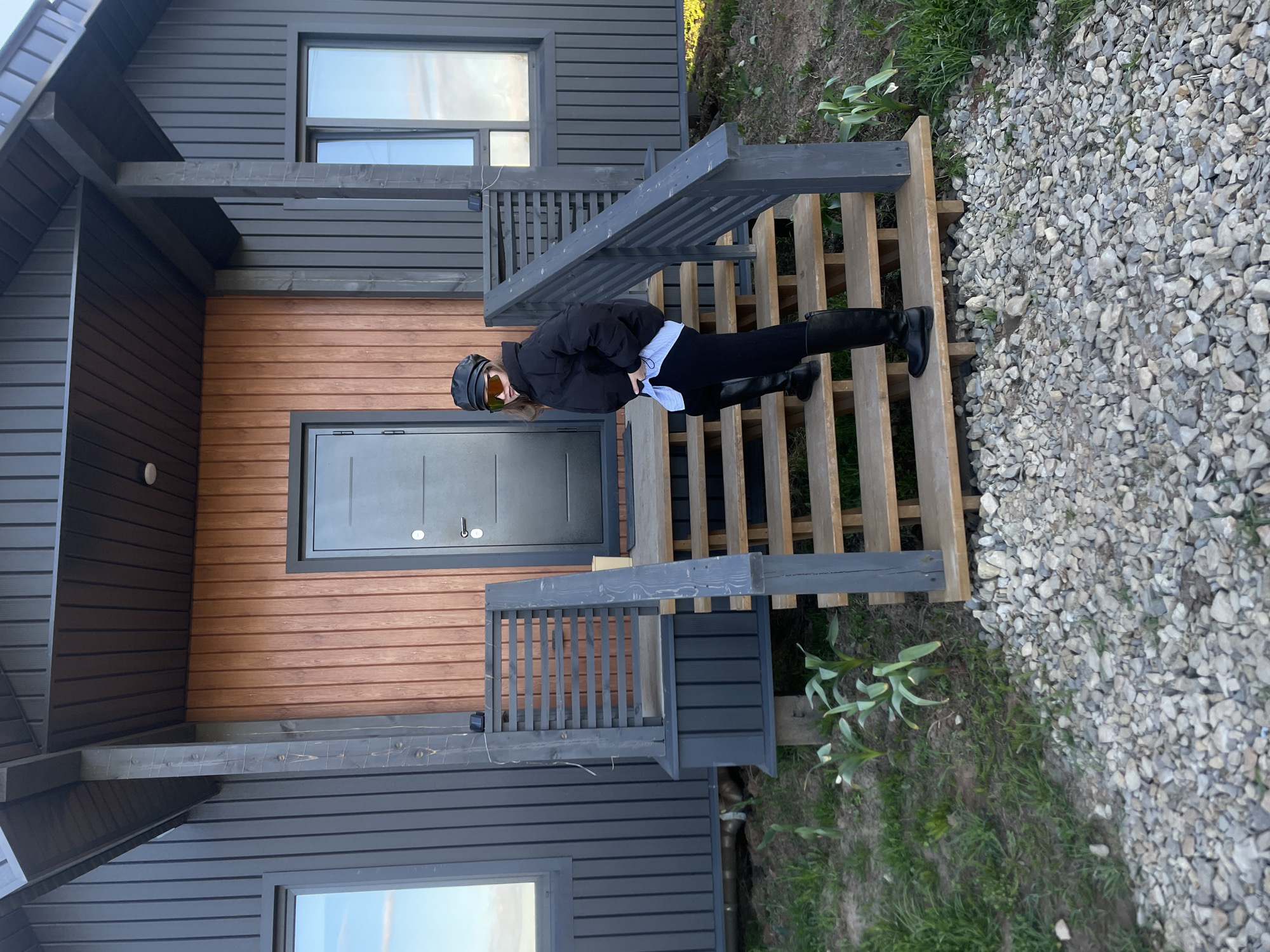 A person in a black jacket, black pants, and black boots stands on a wooden staircase in front of a building. The building has a combination of brown and dark gray wood siding. A gray door is centered on the brown portion of the siding. The stairs are painted a similar dark gray. The background consists of grass, small rocks, and plants. Based on geotags, the location is Подшивалово, Russia.