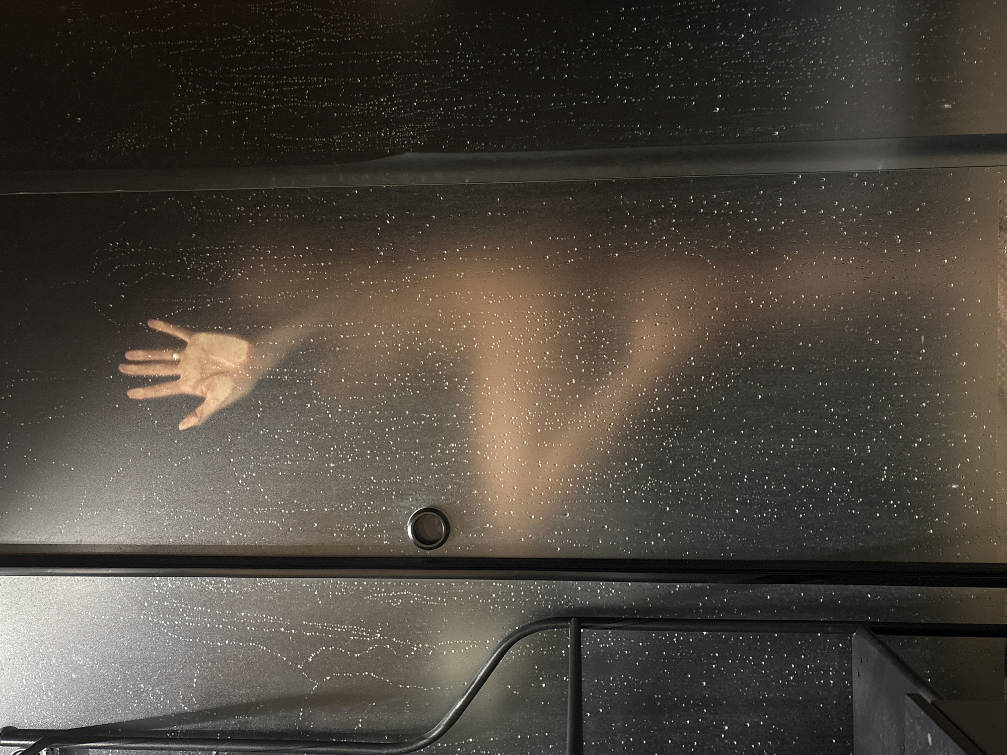 The image captures a human figure, specifically an arm and partial torso, seen indistinctly through a translucent or frosted barrier, likely a shower screen or sauna wall. A person's right hand, wearing a ring on the fourth finger, is pressed firmly against the barrier from the inside, appearing in clearer detail than the rest of the body.
The transparent surface of the barrier is extensively covered with numerous small water droplets, suggesting a steamy or wet environment typical of a bathroom, shower, or sauna. A dark horizontal band runs across the middle of the image, separating the upper and lower sections of the barrier, possibly a structural element of the enclosure. In the lower half of the image, a small circular metal fixture is visible near the center, which could be a handle or part of a drainage system. Towards the bottom right, a dark, curved metal railing or support bar is present.
The scene conveys a sense of privacy and intimacy within an indoor setting. There are no explicit indicators of the time of day or external weather. No visible text is present in the image. The provided location context for this media file is Chaikovsky, Russia.