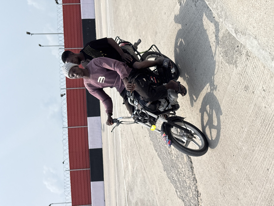 The scene in Igboefon, Nigeria, depicts two males on a black motorcycle on a light grey concrete road surface under bright daylight. The individual in the driver's position wears a light grey knit beanie, a plum-colored long-sleeved sweatshirt with a white 'B' logo, dark trousers, and black flip-flops. He grips the motorcycle's chrome handlebars. Seated directly behind him is a second male, attired in a dark short-sleeved t-shirt with vertical yellow embellishments, dark shorts, and blue and white patterned slides. The motorcycle features silver alloy wheels, a dark front fender with colorful decals, and a yellow rectangular reflective sticker on the front fork. No helmets are worn by either occupant. Distinct shadows are cast on the road. The background consists of a red barrier wall topped by a black and white striped barrier, above which a fence secured with coiled razor wire is visible. Two street light poles stand in the distance against a pale blue, hazy sky.