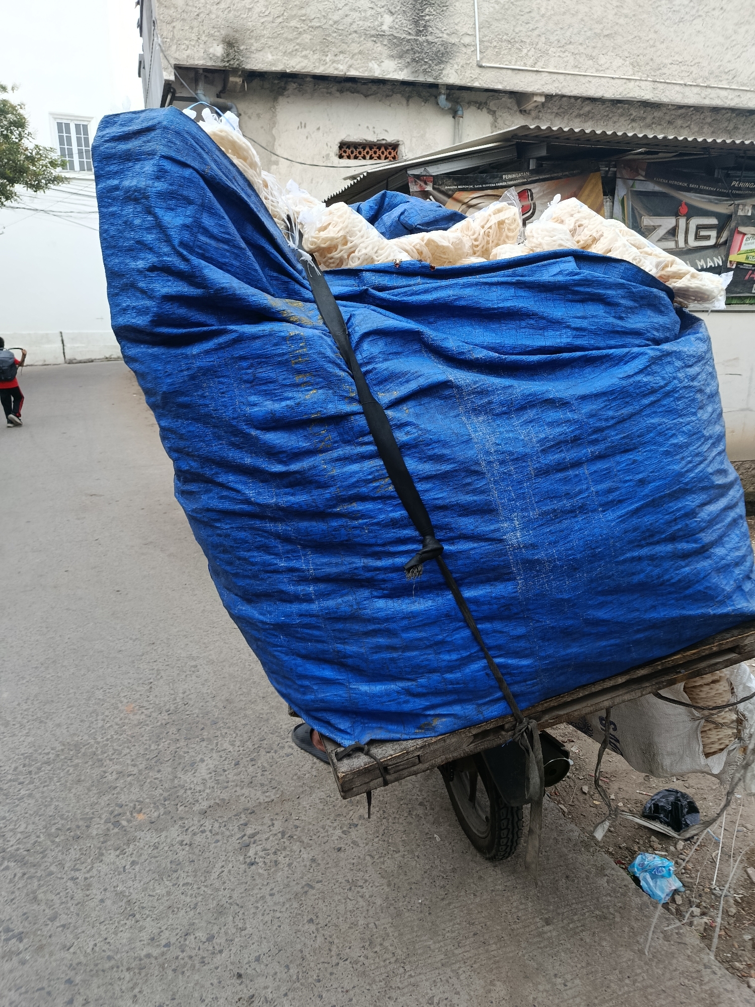 In Balaraja, Indonesia, a large blue tarp is draped over a substantial load, likely food products like crackers or snacks, secured to the back of a motorcycle. The tarp is tied down with a black strap. The motorcycle, partially visible, has a rear wheel and fender. In the background, buildings with grey and white facades line the street. A person in red and black attire, carrying a backpack, walks away from the motorcycle on the asphalt road. Several plastic bags and debris are scattered on the ground next to the motorcycle. A glimpse of advertising banners is visible on the building in the background.