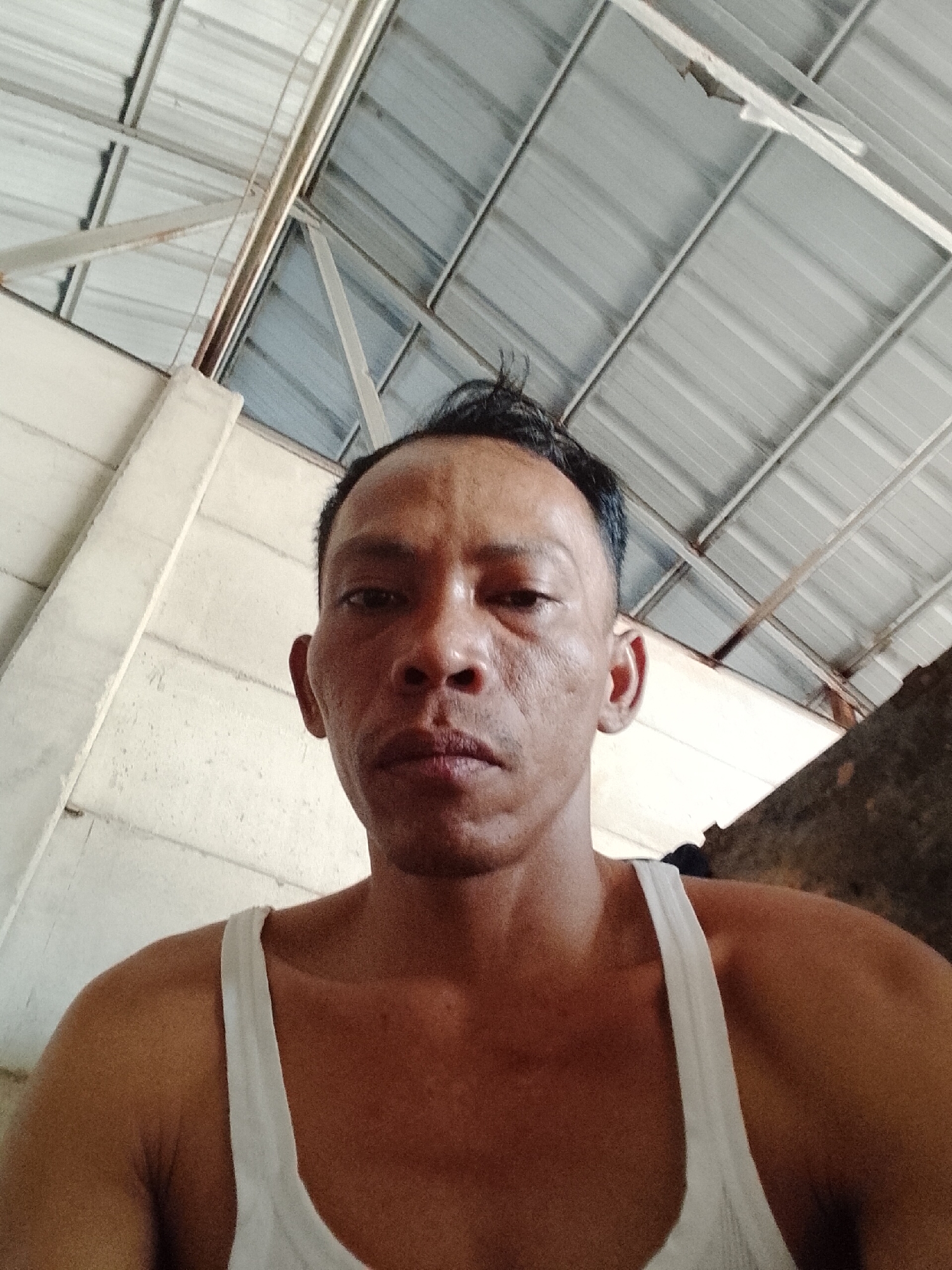 A man wearing a white tank top is pictured from the chest up. He is facing the camera, his expression is neutral. He has short, dark hair. The background shows a corrugated metal ceiling and a white wall. The scene is likely indoors or under a covered structure. There are no discernible landmarks to indicate the specific location within Bukit Ketok, Indonesia.