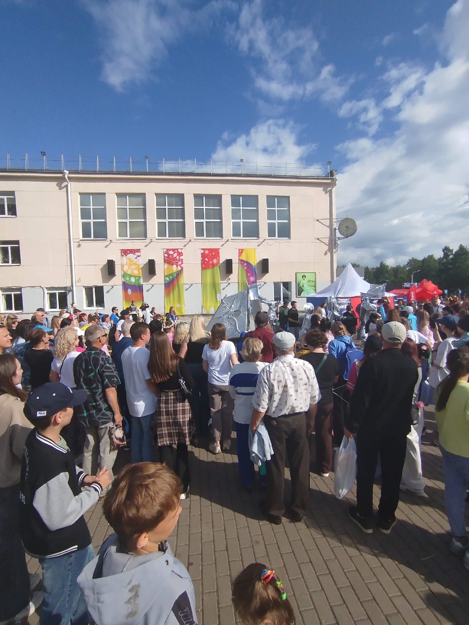 An outdoor event is underway in the city of Chaikovsky, Russia, on a sunny day with scattered clouds. A crowd of people, of varying ages, are gathered in front of a light-colored, multi-story building. The building has several rows of windows and is decorated with colorful banners featuring abstract designs. A clock is mounted on the exterior wall of the building.
In the foreground, a young boy wearing a blue baseball cap and a black and grey jacket is looking to his right. Another young boy, with brown hair, is in the lower right corner of the frame, facing away from the camera. A woman with blonde hair is also visible.
In the middle ground, the crowd is dense. Some individuals are dressed in casual attire, while others appear to be performers in elaborate silver costumes, possibly representing robots or futuristic characters. There are also white tents and red inflatable structures in the background, suggesting a fair or festival. The ground is paved with interlocking bricks. A satellite dish is visible on the roof of the building.