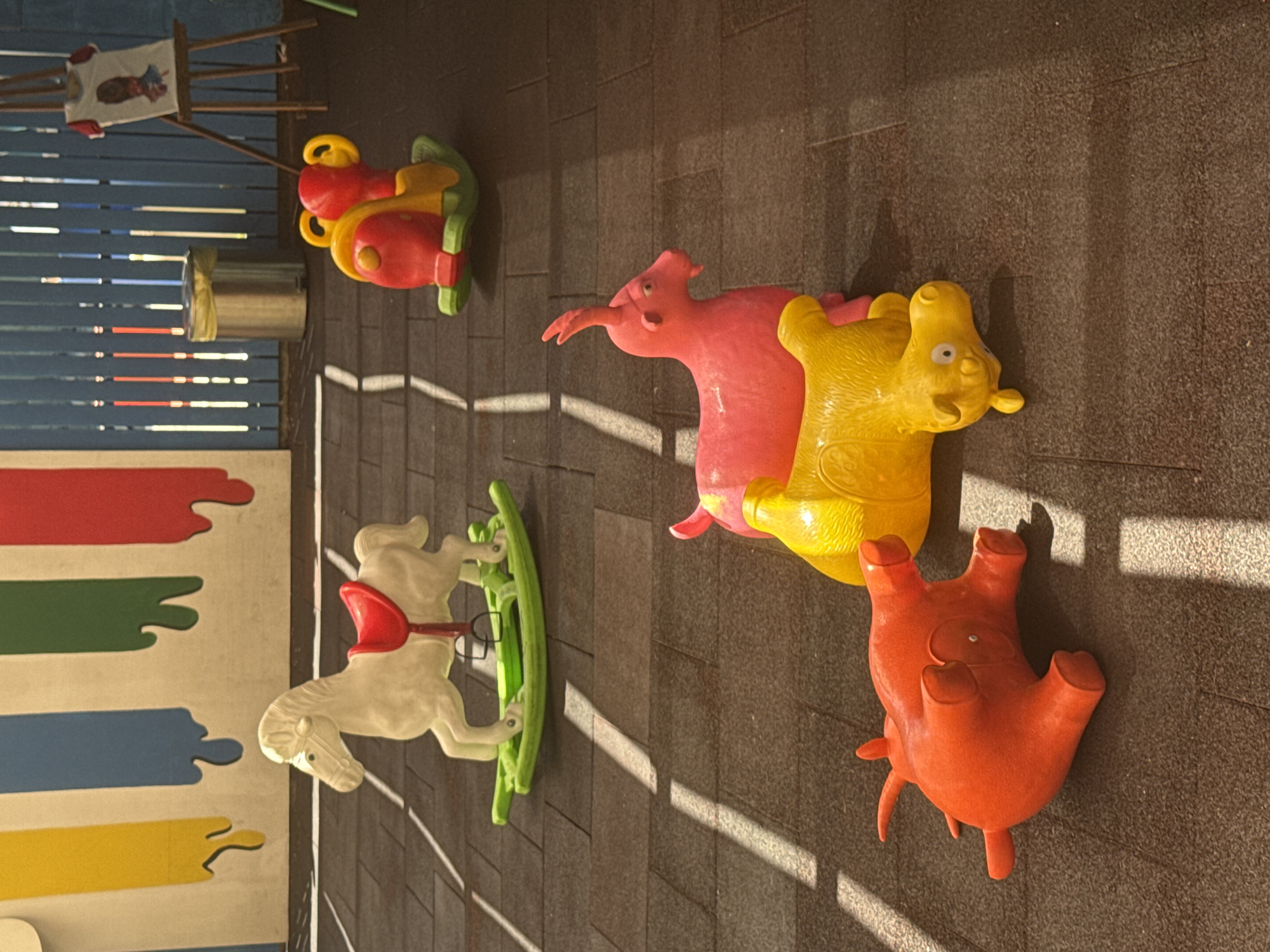The image depicts a children's play area. In the foreground, a bright orange inflatable toy animal, possibly a rhino or hippopotamus, lies on its back on a textured, dark brown surface. Adjacent to it, a yellow inflatable toy animal, also appearing to be a hippopotamus, is positioned on its side. Behind these, a pink inflatable toy resembling a goat with a horn is standing. Further in the background, a white rocking horse with a red saddle is on a green base. To the right of the rocking horse, a red and yellow plastic toy with large handles is on a green base. In the far background, a metal trash can with a yellow lid is visible, along with a wooden easel displaying a framed drawing of a person. The background wall features colorful vertical stripes in yellow, blue, green, and red. The lighting suggests the scene is outdoors, with shadows cast on the ground by sunlight. The exact location in Aksu, Turkey, cannot be determined from the image alone.