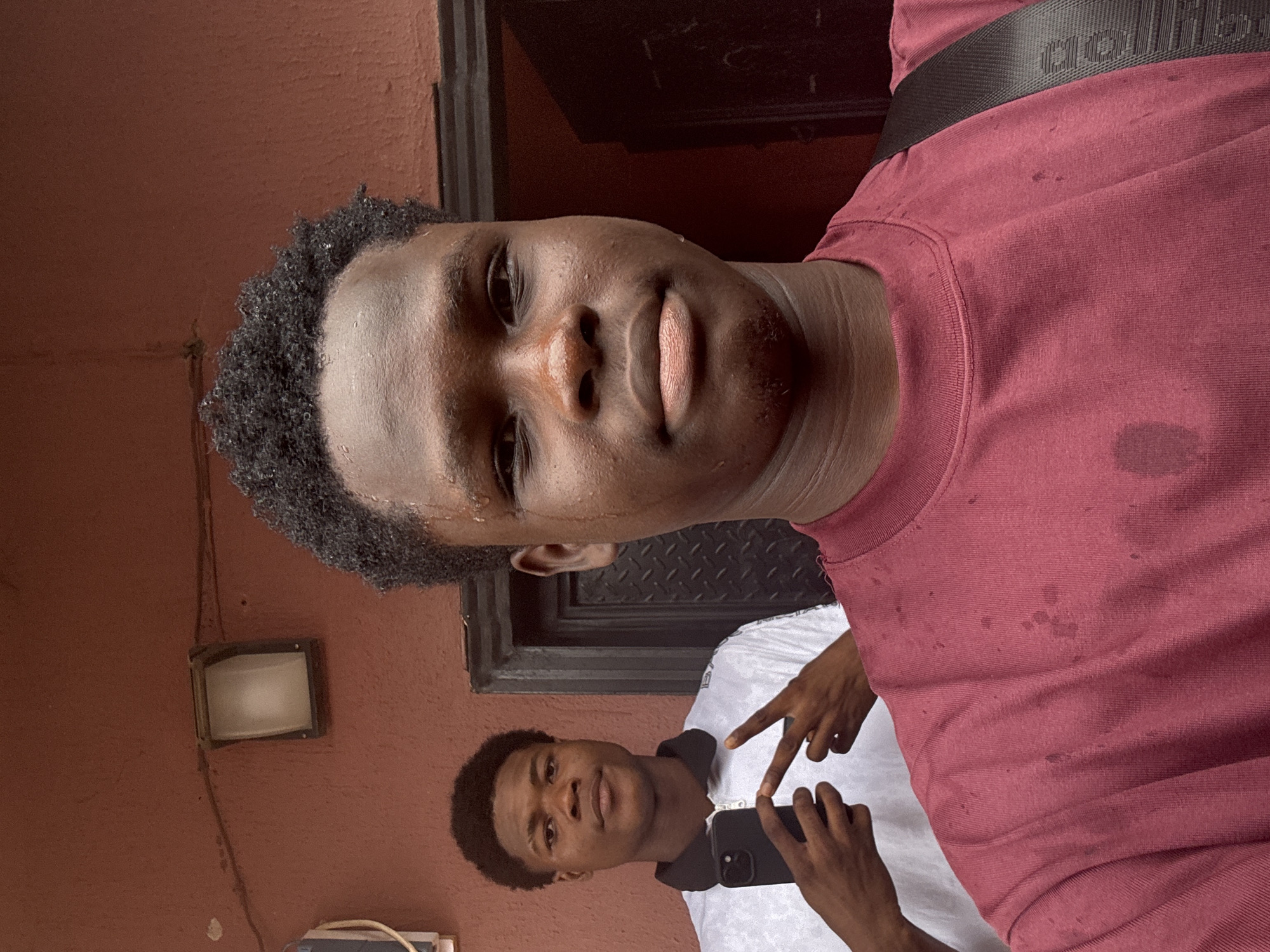 This image, captured in Ihiagwa, Nigeria, features two young Black males in an outdoor setting. The foreground subject, facing the viewer directly, has short, dark, curly hair with visible moisture droplets and wears a maroon crew-neck t-shirt displaying multiple darker spots consistent with sweat or water. A black strap, consistent with a bag or backpack, crosses his left shoulder (viewer's right). The second subject, a young Black male positioned behind and to the left, also faces the viewer. He is dressed in a white collared polo shirt with a black collar and zipper, holding a black smartphone in his left hand and forming a V-sign with his right. The background features a reddish-brown, textured wall. An outdoor light fixture with a rectangular frosted white cover and dark frame is mounted on the upper left, above which a black electrical wire runs horizontally. A dark, textured door frame and a section of a dark door are visible to the right. A portion of a light-colored utility box is visible on the far left.
