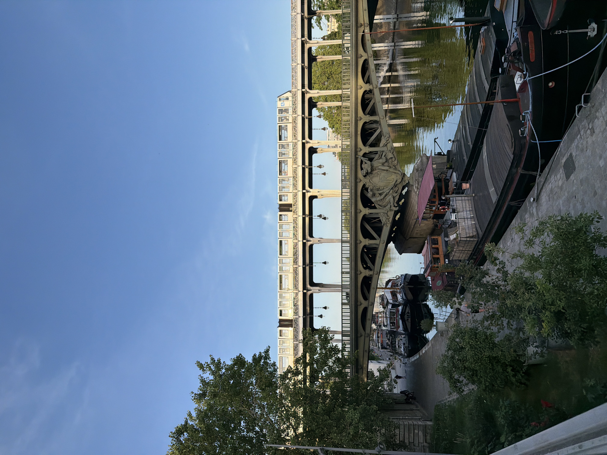 This picture, taken in Paris, France, depicts a section of the Pont de Bir-Hakeim. The image appears to have been rotated 90 degrees counterclockwise. The bridge spans the Seine River, and a pedestrian walkway and elevated metro line can be seen. A number of houseboats are tied along the bank. A blue sky forms the backdrop. A tree grows in the foreground.