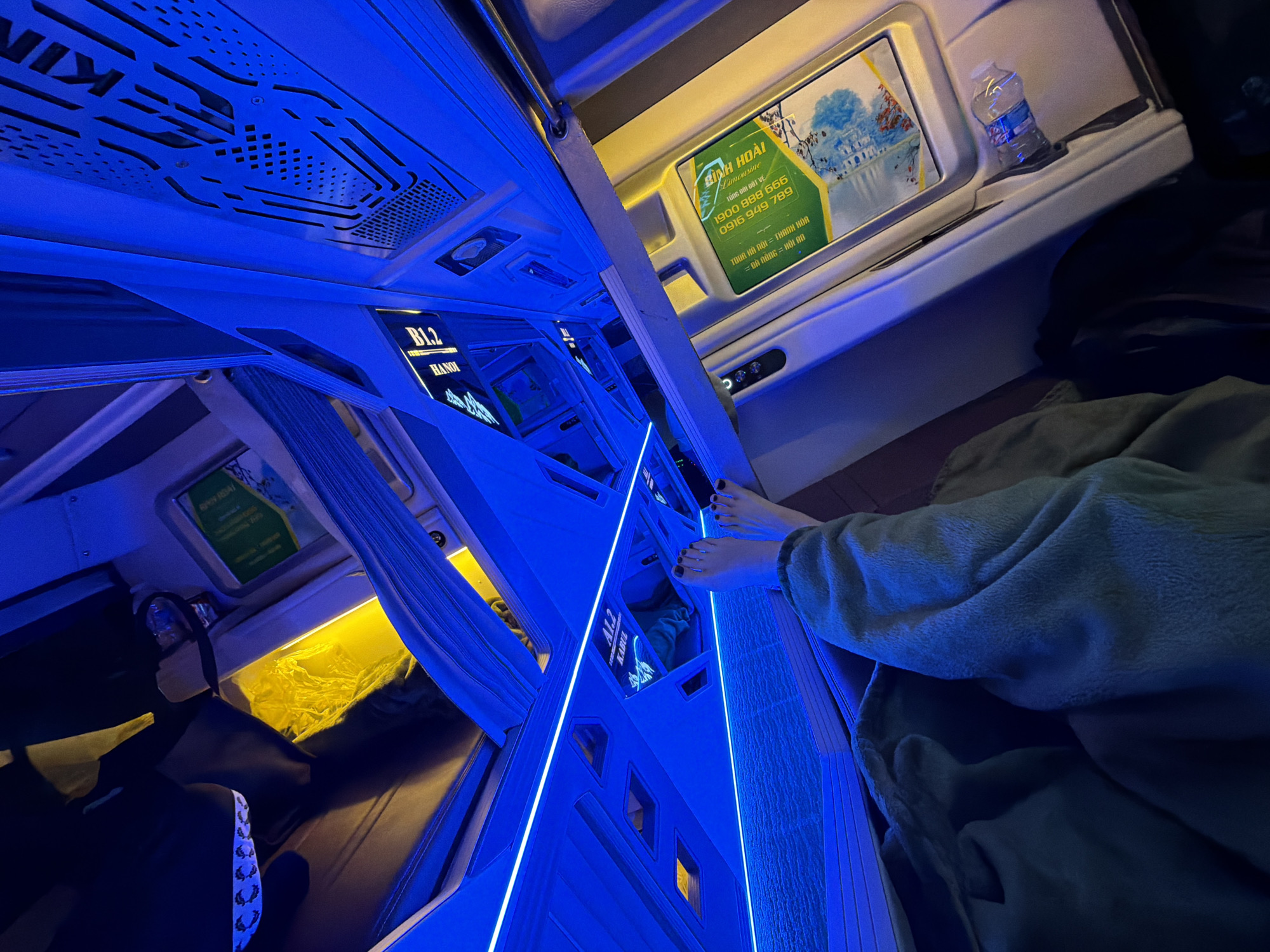 The image displays the interior of a modern sleeper bus, characterized by capsule-style bunks illuminated predominantly by cool blue LED strip lighting. A person's bare feet, with dark painted toenails, extend from one of the upper bunks, partially covered by a soft, fuzzy green-grey blanket or robe, suggesting relaxation or travel.
In the background, a warm yellow light emanates from inside another bunk. A small plastic water bottle is placed in a holder on a shelf near a display screen. Visible text on various displays includes "B1.2 HANOI" and "A1.2 KABUL," likely denoting bunk identifiers or destinations.
A prominent green and yellow advertisement for "BÌNH HOÀI Limousine" provides contact numbers ("1900 888 666", "0916 949 789") and lists routes: "TUYẾN HÀ NỘI = THANH HÓA" and "= ĐÀ NẴNG = HỘI AN," confirming the location context of Đà Nẵng, Vietnam. The ad also features a scenic graphic. Partial text "KIN" and "RUN ROAI" are visible on the ceiling and upper structures of the bus interior. The low light conditions, coupled with the specialized bunks and route information, indicate a long-distance, possibly overnight, journey.
