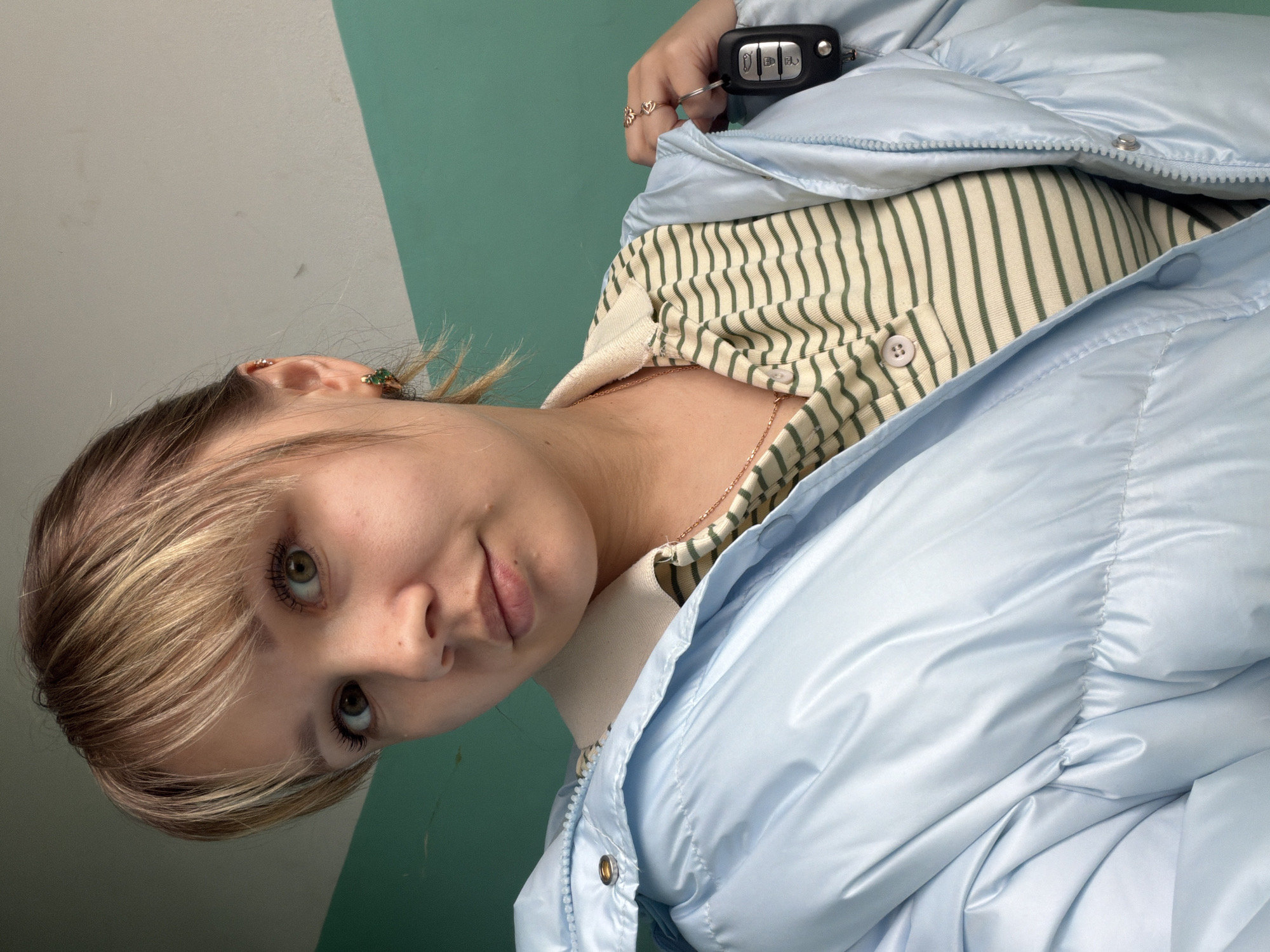 This portrait-oriented image, rotated 90 degrees counter-clockwise, features a young individual, likely female, positioned in the foreground. She is looking directly at the camera with light green or hazel eyes and a subtle, almost neutral expression. Her hair is light-colored with blonde highlights, styled with bangs across her forehead.
She is dressed in a shiny, light blue puffer jacket, partially unzipped, revealing a cream-colored long-sleeved shirt with thin green horizontal stripes underneath. A delicate gold-toned chain necklace is visible around her neck. In her right hand, she holds a black car key fob with silver buttons. Visible text and icons on the fob include "LOCK," "UNLOCK," a trunk icon, and a car icon. She wears multiple rings on this hand, including one with small heart shapes. A green, possibly jeweled, earring is visible in her left ear.
The background is a wall divided into two sections by an angled line: a light grey or off-white on the left and a teal green on the right. The lighting is bright and even, without harsh shadows, suggesting an indoor or sheltered outdoor setting. The overall mood is calm and direct.