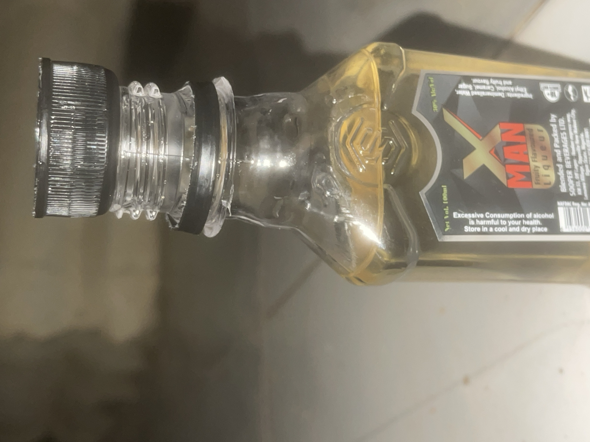 The image shows a close-up of a small glass bottle containing a yellow liquid. The bottle has a black plastic screw-on cap and a clear plastic neck with ribbed detailing. The label on the bottle is predominantly black with red and yellow lettering. The brand name appears to be "X MAN" with the tagline "Fruity Flavoured Liqueur." Other text on the label includes "Net Vol. 100ml," "30% Alc/Vol," "Ingredients: Demineralised Water, Ethyl Alcohol, Caramel, Sugar and Fruity flavour," and "Blended and Packed by SOOPER BEVERAGES LTD." There is also a warning about the excessive consumption of alcohol. The background is out of focus but appears to be a neutral-colored, possibly tiled or concrete floor. There are no people or discernible activities depicted in the image. The image does not provide any information about the location in the city of Howayi, Nigeria, or any other specific location.
