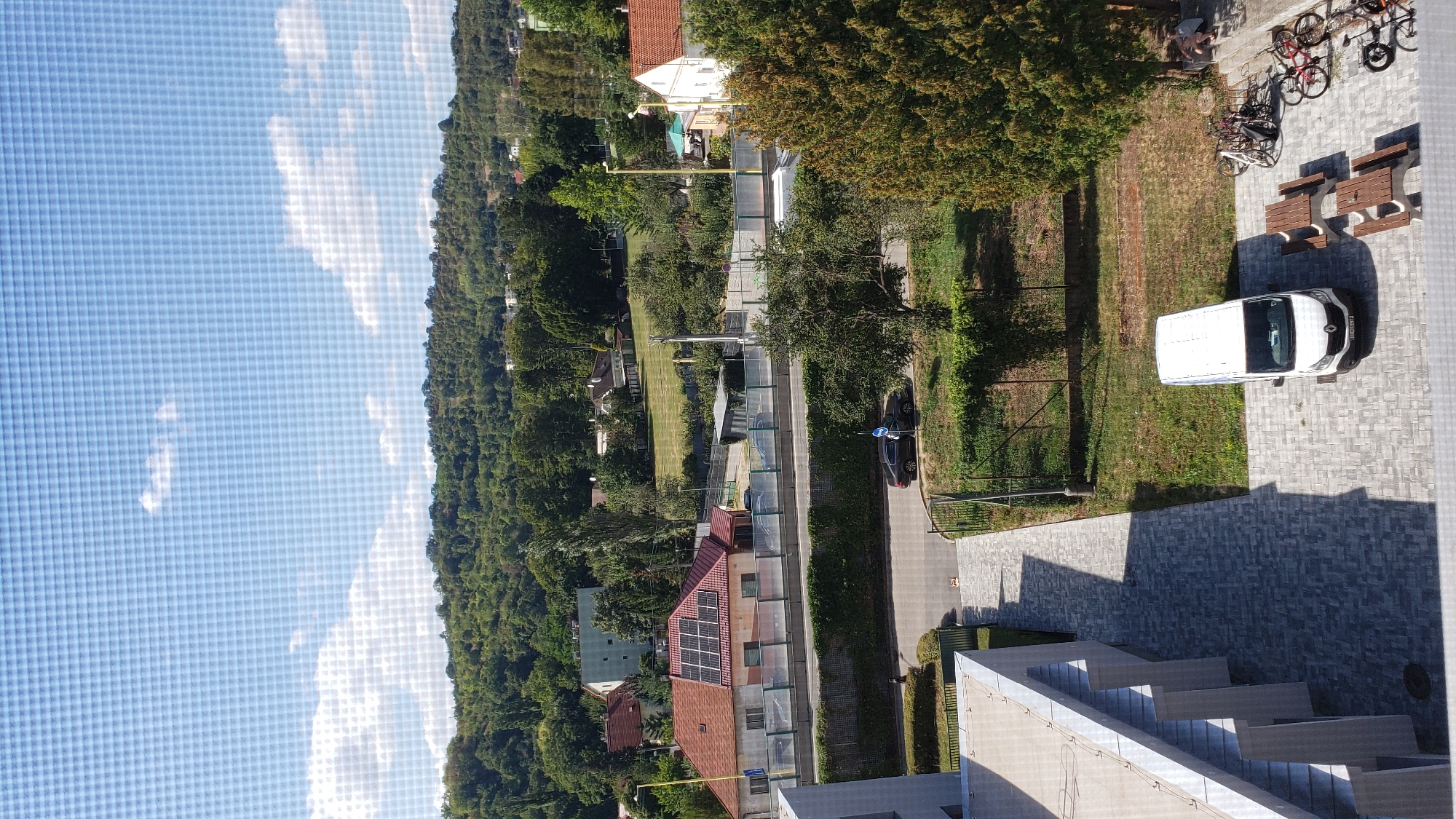 The image captures a daytime view of a residential area, likely in Prešov, Slovakia, given the context of the query. A modern building with a light-colored facade and visible support columns is in the foreground on the left. Adjacent to it is a paved walkway with a patterned gray stone surface, leading towards a street. A white van is parked on the paved area, and several picnic tables are set up nearby. To the right of the van, a cluster of bicycles is parked, some of which appear to be chained together. A large evergreen tree stands to the right of the paved area, with its branches extending into the frame. Further in the background, a street runs horizontally, with a dark-colored car parked on it. Across the street, there is a house with a red roof and solar panels, as well as other residential buildings and lush green trees lining a hillside. The sky above is blue with scattered white clouds. A person can be seen sitting on a step near the evergreen tree.