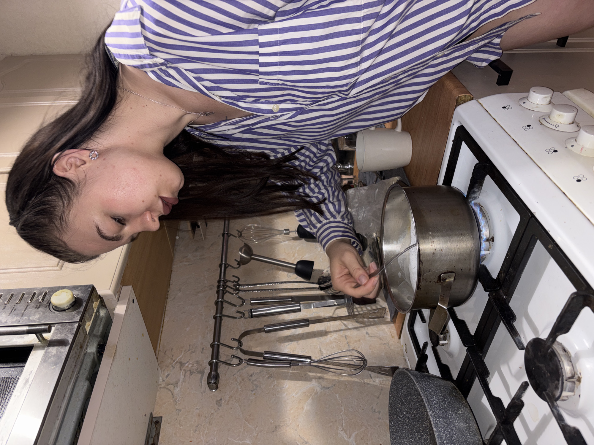 A woman with long, dark hair, wearing a blue and white striped shirt and jewelry, stands in a kitchen. She is looking down at a pot on a gas stovetop, stirring its contents with a utensil. A rack of kitchen tools hangs beside the stovetop. A white mug and a frying pan are also visible on the countertop. The image does not provide sufficient information to confirm the location in Perm, Russia, or any other city.