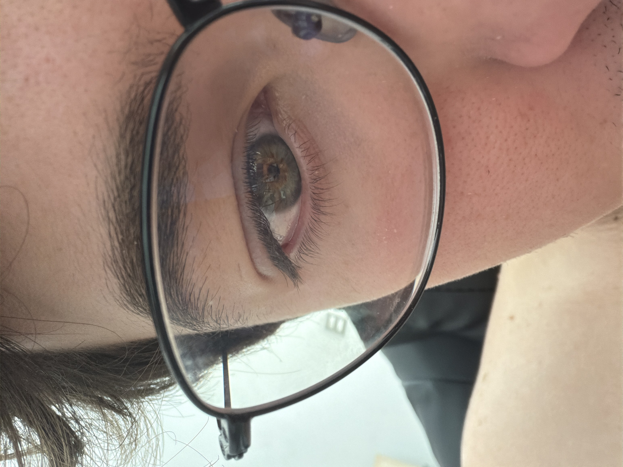 A close-up image focuses on a person's right eye, magnified through a lens of eyeglasses. The iris is visible, revealing green and brown tones. The eyebrow is dark and thick. The surrounding skin has fine lines and pores visible. The frame of the glasses is black. The background is indistinct, but appears to be a light-colored wall. It is difficult to identify the specific location within San Andrés Cholula, Mexico based solely on this image.