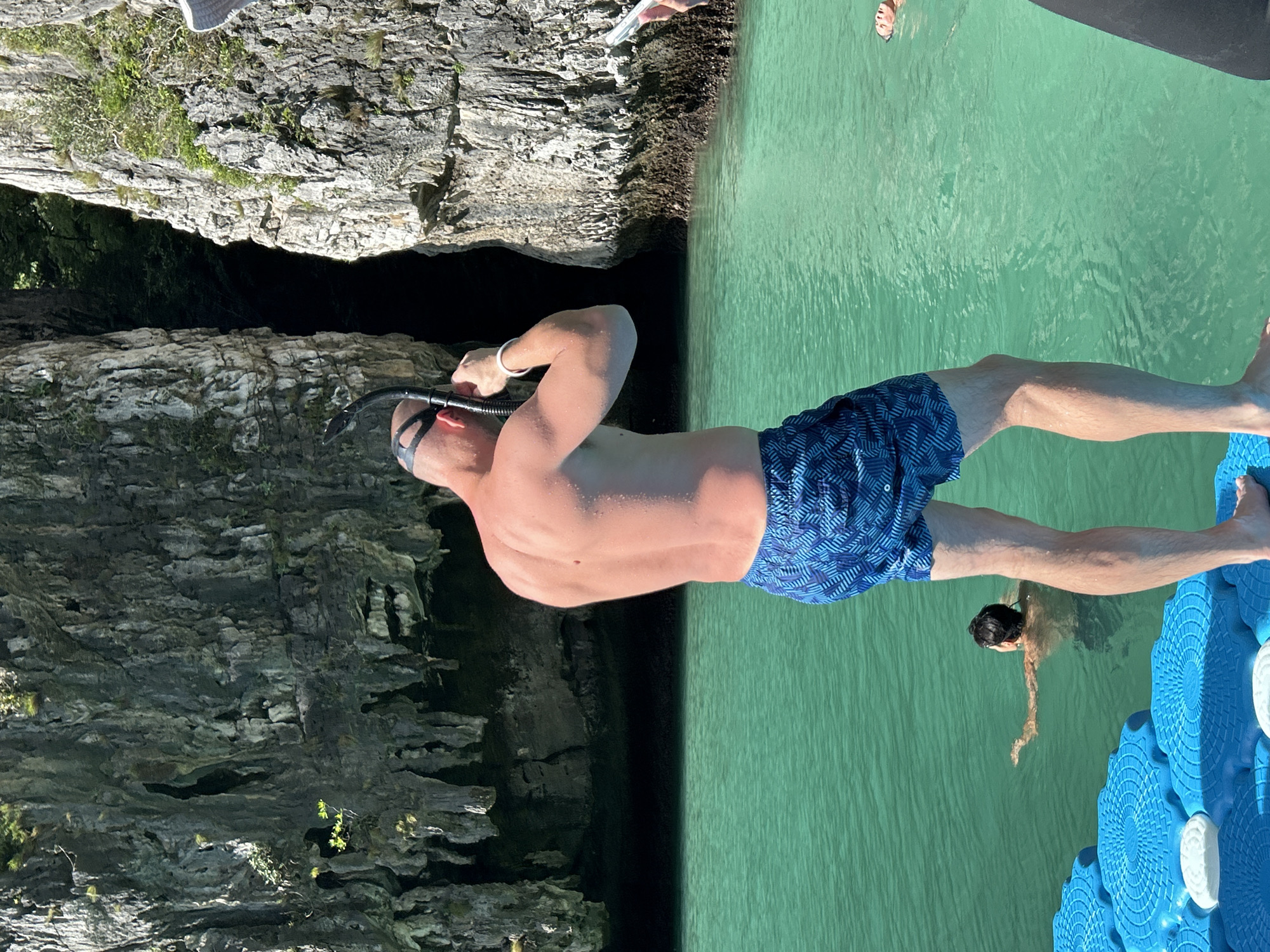 The image depicts a man in blue patterned swim trunks and a snorkeling mask preparing to enter the water. He stands on a blue floating platform, facing away from the camera towards a narrow opening in a limestone cliff face. The water is a vibrant turquoise color. In the background, two other individuals are visible swimming in the water. The environment suggests a tropical location, likely within the karst landscape of Ao Nang, Thailand, known for its dramatic cliffs and clear waters.