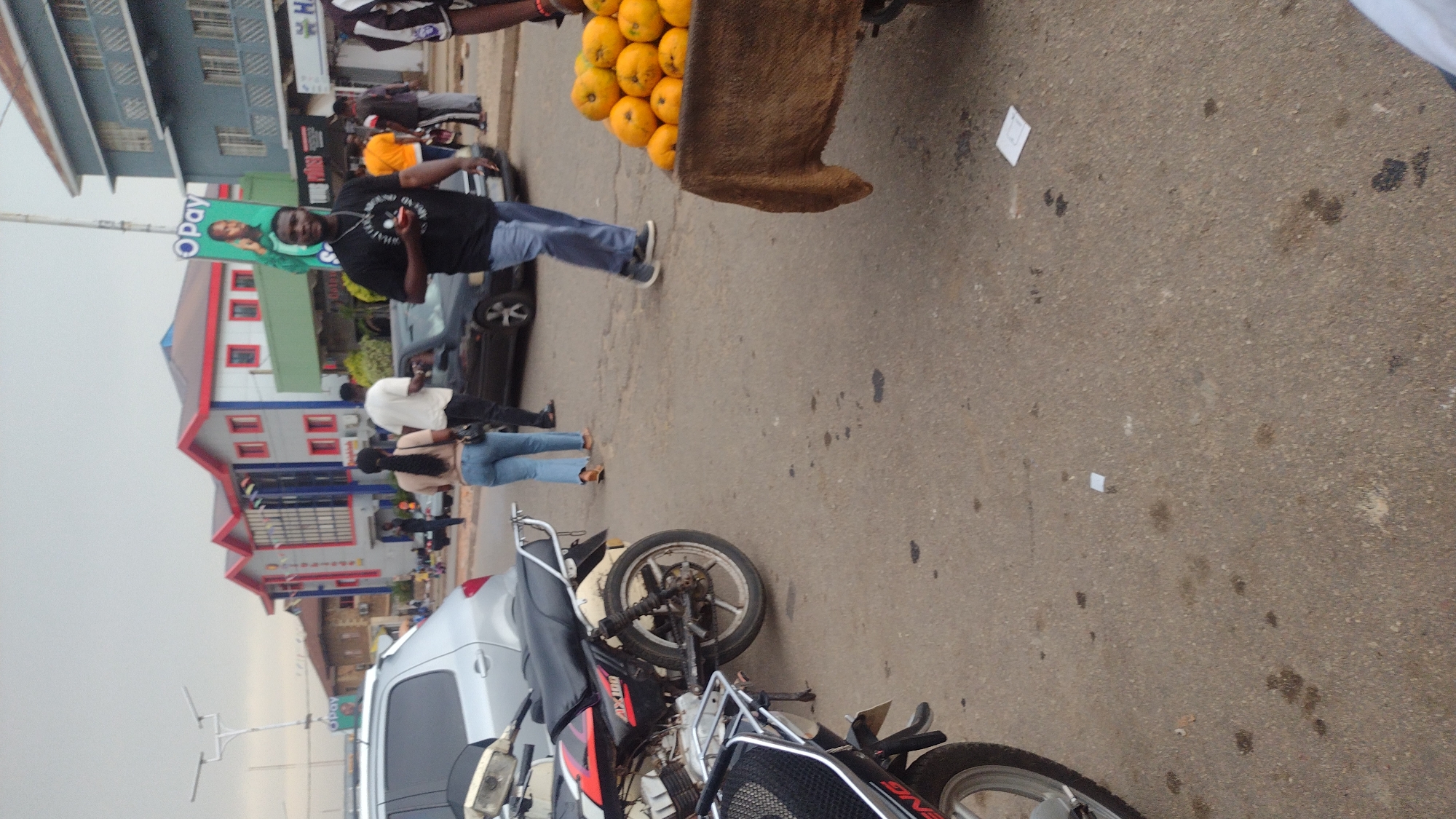 The image captures a street scene in Jos, Nigeria. In the foreground, a motorcycle is parked with a silver car behind it. Further up the frame, a vendor is pushing a cart laden with what appears to be oranges. Several people are seen walking around, including a man taking a picture with his phone. Shops and buildings are visible in the background with signs on some. The sky is overcast. The street is paved with dark spots and some litter.