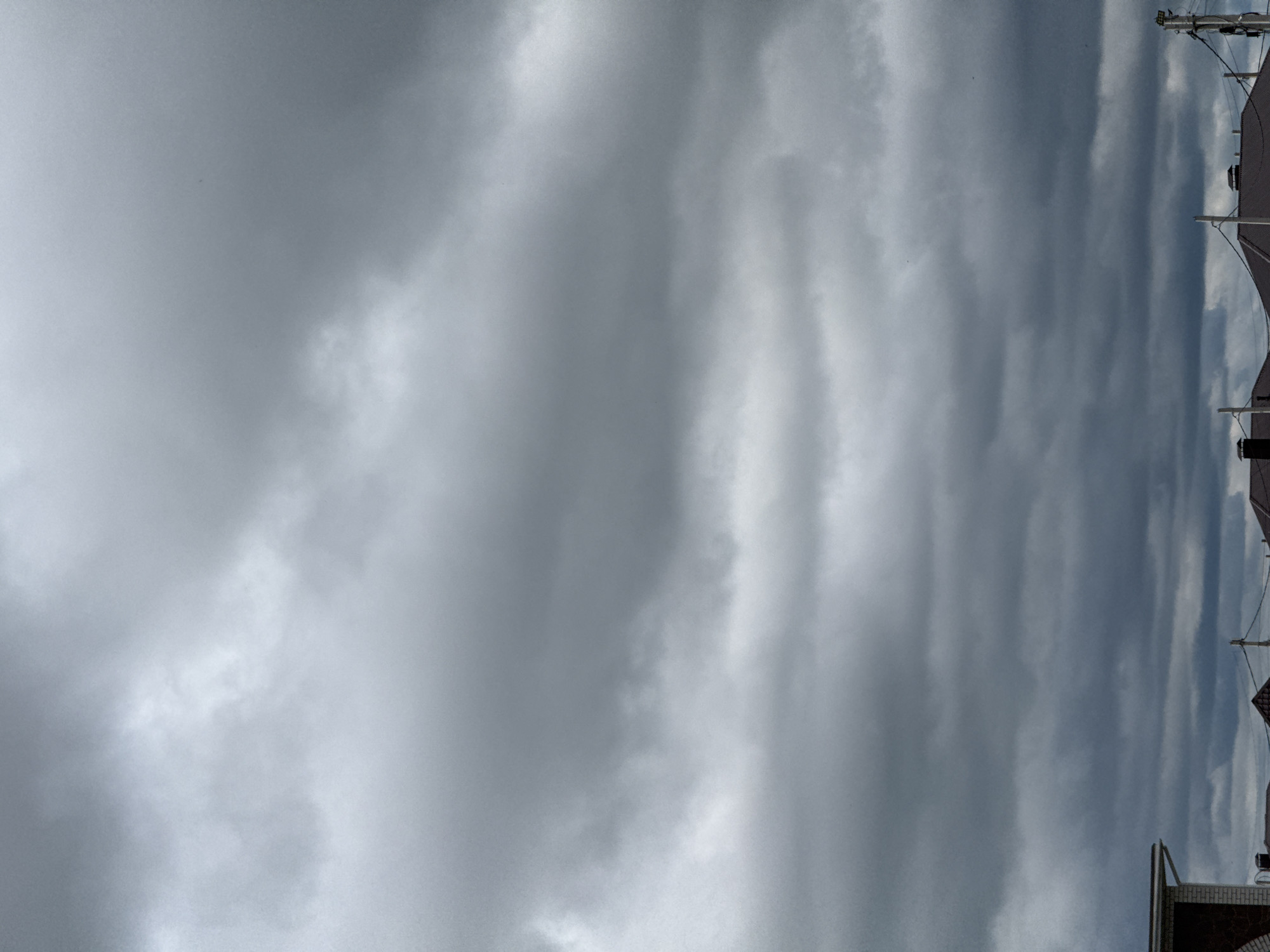 The image shows a cloudy sky above a residential area, likely in Murom, Russia, given the provided context. The sky is filled with dense, gray clouds that suggest an overcast or impending rainy weather. Below the sky, the roofs of several houses are visible, some with chimneys. Power lines and utility poles are also present, indicating an urban or suburban setting. The overall scene is muted in color due to the heavy cloud cover, with the predominant tones being various shades of gray and dark brown from the roofing materials. No people are visible in the frame. The composition is dominated by the sky, with the rooftops forming a lower horizontal band across the bottom of the image.