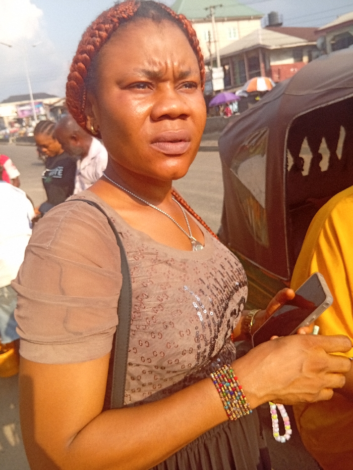 The scene is an outdoor urban environment in Ura Nta Umuarandu, Nigeria, observed during daylight. The primary subject is an adult female with brown skin and reddish-brown braided hair styled in cornrows, depicted from the chest up, positioned slightly left of center. She wears a light brown or taupe-colored top featuring sheer, ruched sleeves and a sequined pattern resembling "GG" lettering on the torso. A thin silver-colored chain necklace with a small pendant is visible. Her left wrist displays multiple colorful beaded bracelets, and a white and pastel beaded bracelet hangs from her left hand. Her facial expression is serious, with a furrowed brow, as she looks towards the right, off-frame.
To her immediate right, partially visible, is an individual wearing a yellow shirt, holding a black-screened smartphone in their right hand. The primary subject's left hand is positioned near this smartphone. In the mid-ground, behind the primary subject and to her left, two blurred individuals are visible: one female with braided hair wearing a dark shirt, and one male in a light-colored shirt. To the primary subject's right, a brown three-wheeled vehicle, resembling an auto-rickshaw, is partially visible. The background features various buildings, including a light-colored multi-story structure with a dark roof, a utility pole, and two market umbrellas—one orange and white, the other purple. The ground appears to be paved.
