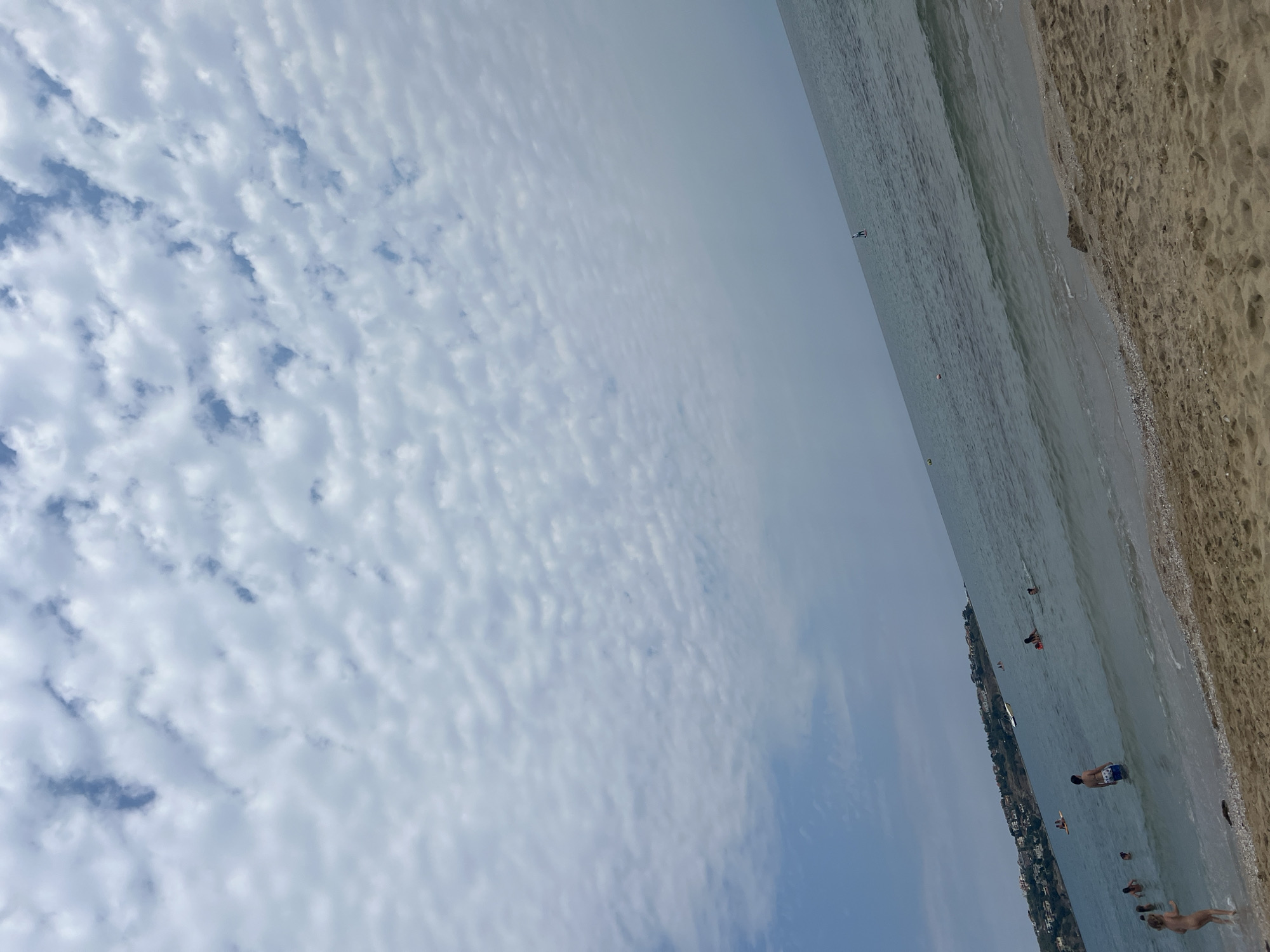 The image depicts a beach scene in Sozopol, Bulgaria, under a cloudy sky. The foreground consists of sandy beach with some pebbles. The sea stretches into the distance, with small waves lapping at the shore. Several people are visible in the water, swimming, wading, and some standing near the shore. One person is on a paddleboard in the middle distance. In the background, a coastline with buildings and trees is visible. The overall impression is of a leisurely day at the beach.