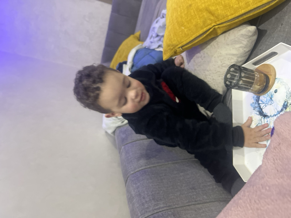 A child in a black tracksuit sits on a gray couch with yellow and white pillows. The child is reaching for a white tray holding a clear container with a wooden base and a floral design. The child appears to be trying to grab or move the tray. A pink blanket is partially visible in the foreground. The wall is light and plain.