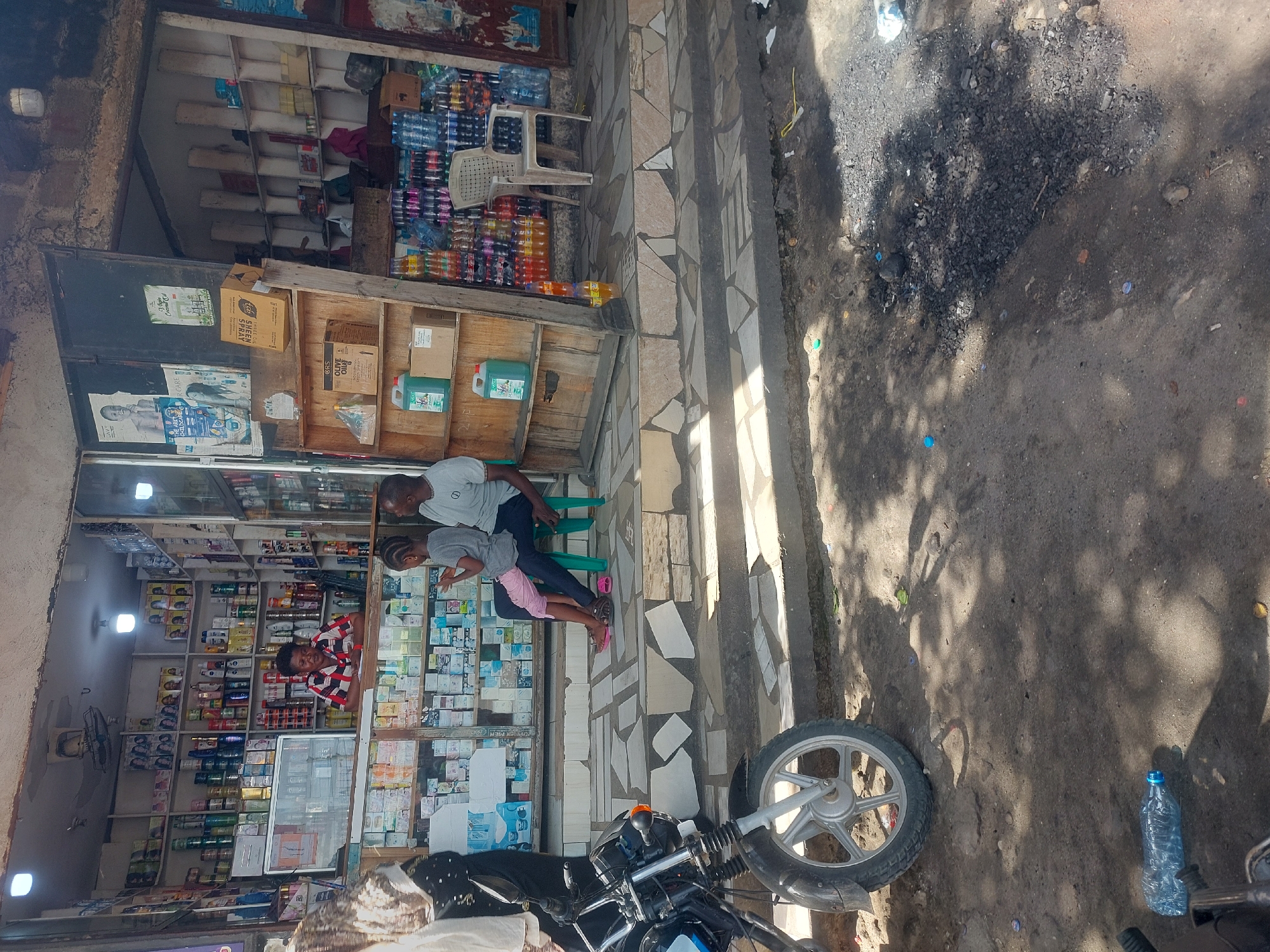 A street scene in Dar es-Salaam, Tanzania shows a storefront with various items for sale. A man and child are standing outside the storefront, interacting with someone inside. A motorcycle is parked in front of the store. A variety of goods are visible on the shelves inside the shop. The environment outside the store includes a dirt and paved sidewalk area. A plastic water bottle lays on the ground.