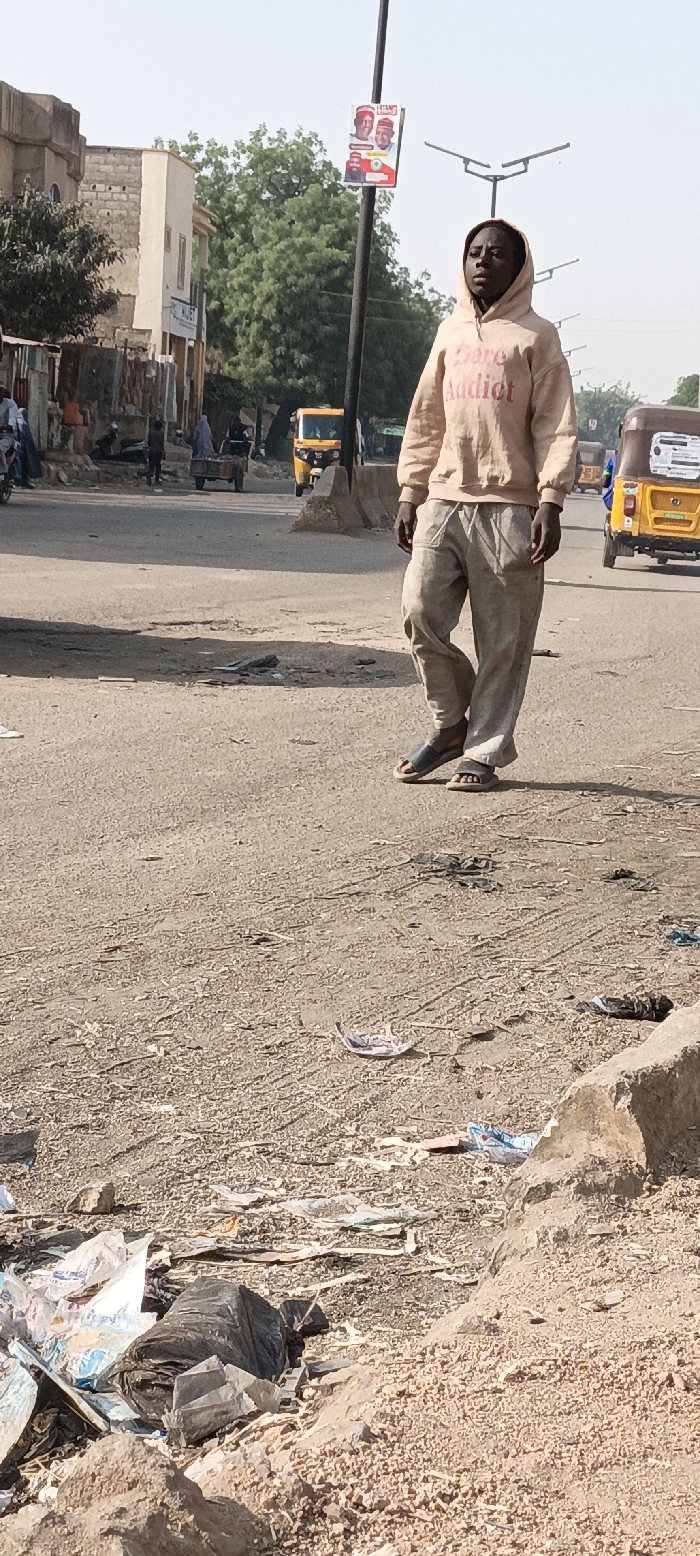 An outdoor street scene in Kaura Goje, Nigeria, is depicted under clear daylight. A dark-skinned individual, appearing male, stands in the center-right foreground, facing slightly left with a neutral expression. He wears a light peach-colored hooded sweatshirt with "Dare Addict" printed in reddish-brown text, loose light grey pants, and dark grey open-toed sandals.
The immediate foreground consists of rough, unpaved earth covered extensively with litter, including crumpled plastic bags (one black, one white/blue) and paper debris. Beyond this, a paved road extends into the midground, featuring concrete median barriers.
On the left side of the road, several indistinct figures are visible, along with a yellow three-wheeled auto-rickshaw. A person in a light-colored full-body garment pushes a two-wheeled cart along the road. On the far right, another yellow three-wheeled auto-rickshaw is present.
In the background, a line of buildings constructed from light-colored blocks and painted facades is visible on the left, interspersed with green trees. A dark street light pole stands to the left of the central individual, bearing a rectangular political poster featuring two smiling men in red caps and a red, green, and white national emblem. Additional streetlights with three-pronged fixtures are seen further down the road. The sky is clear and light blue.