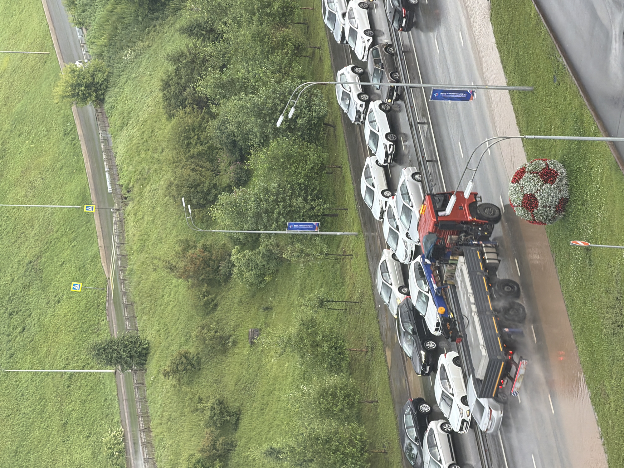 This is an aerial, vertically oriented photograph of a traffic jam on a wet asphalt road. The road is lined with lush green grass and trees on the left side, and a grass verge on the right. Several cars are stopped on the road, most of them white, with a few black cars interspersed. A large red truck with a flatbed trailer is also part of the traffic jam. The ground is wet, suggesting recent rain, and there are visible water splashes around the tires of the vehicles.
In the distance, there is a decorative spherical object covered in red and white flowers, resembling a soccer ball. There are also streetlights and traffic signs visible. One sign on the road is blue with white text that is partially obscured but appears to read "2025". Another yellow sign with a pedestrian symbol is visible on the grassy area to the left of the road. The scene appears to be in an urban or suburban setting, with the presence of infrastructure like roads, streetlights, and signs. The overcast sky and wet road indicate it is likely a cloudy or rainy day. There are no discernible people in the image.