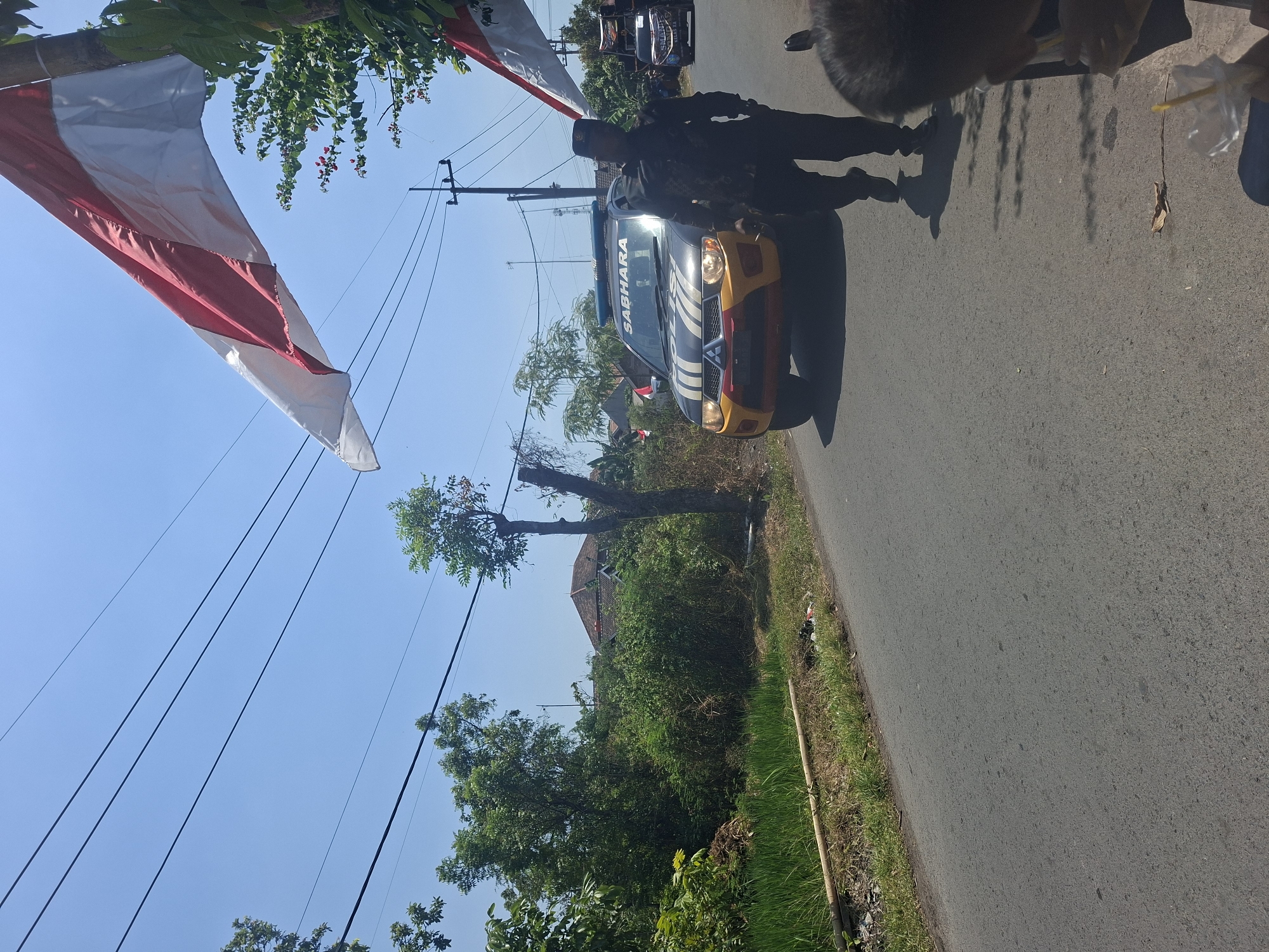 In Tongaswetan, Indonesia, under a clear blue sky, an Indonesian flag flutters from a pole, partially obscuring a view of telephone wires overhead. A dark-colored Mitsubishi Triton pickup truck, marked with "SABHARA" on its windshield and police insignia, drives down a paved road. A uniformed officer stands beside the truck, facing the camera. The truck's lights are illuminated. To the left of the road, lush green vegetation and a rice paddy are visible. In the distance, houses with terracotta roofs are nestled amongst trees. Another police vehicle, a similar pickup truck, is seen further down the road, also with police markings. The foreground shows a person's head and shoulder in the bottom right corner of the frame, with a plastic cup and straw. The scene appears to be a sunny day, possibly during a parade or an official event.