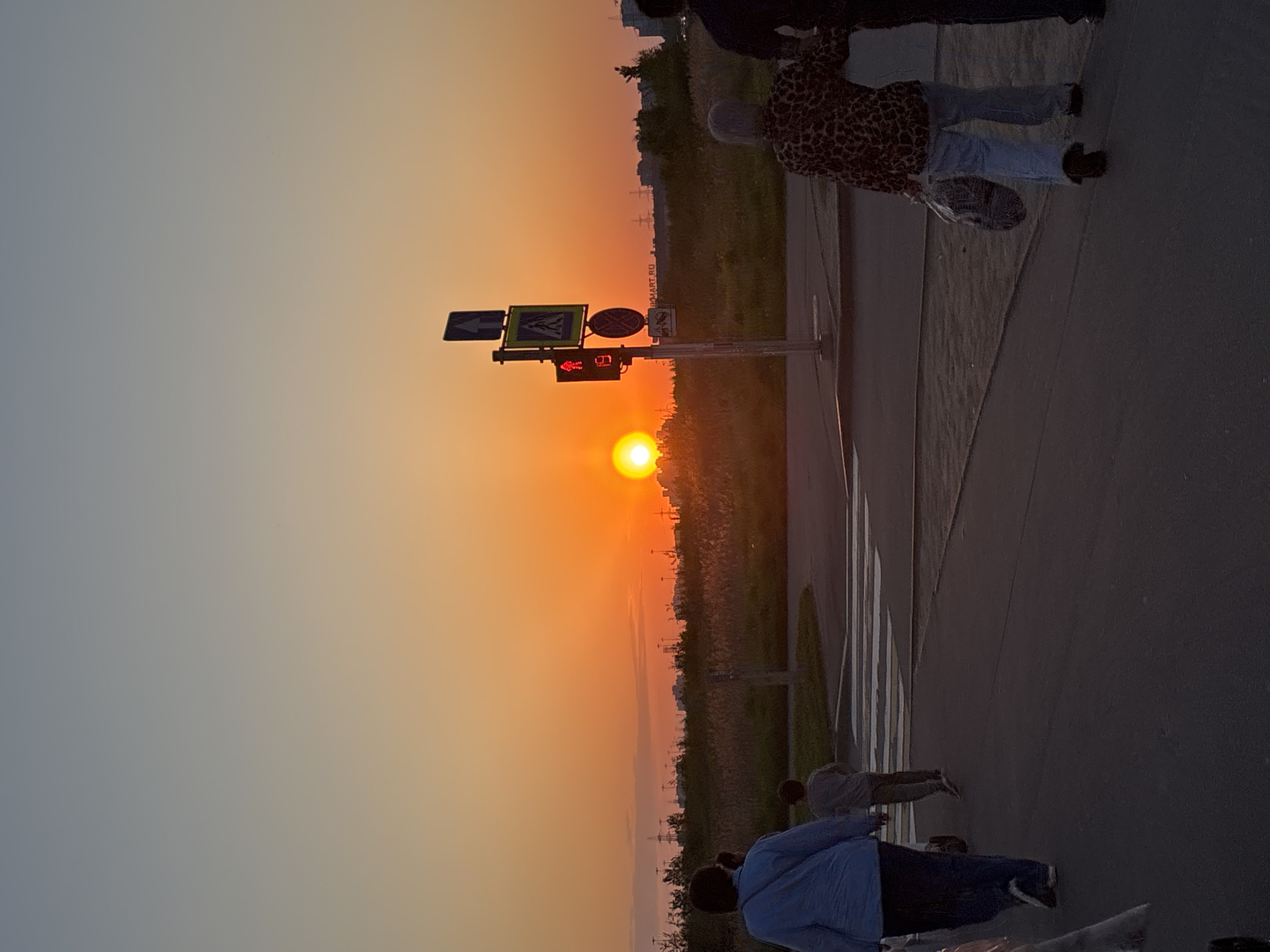 The image depicts a sunset in Murino, Russia. The sun is low on the horizon, appearing orange, and situated above a pedestrian crossing with a traffic light displaying a red signal. A group of people are visible on the sidewalk near the crossing, and there are buildings and vegetation in the background. The overall scene conveys an urban environment during the evening.