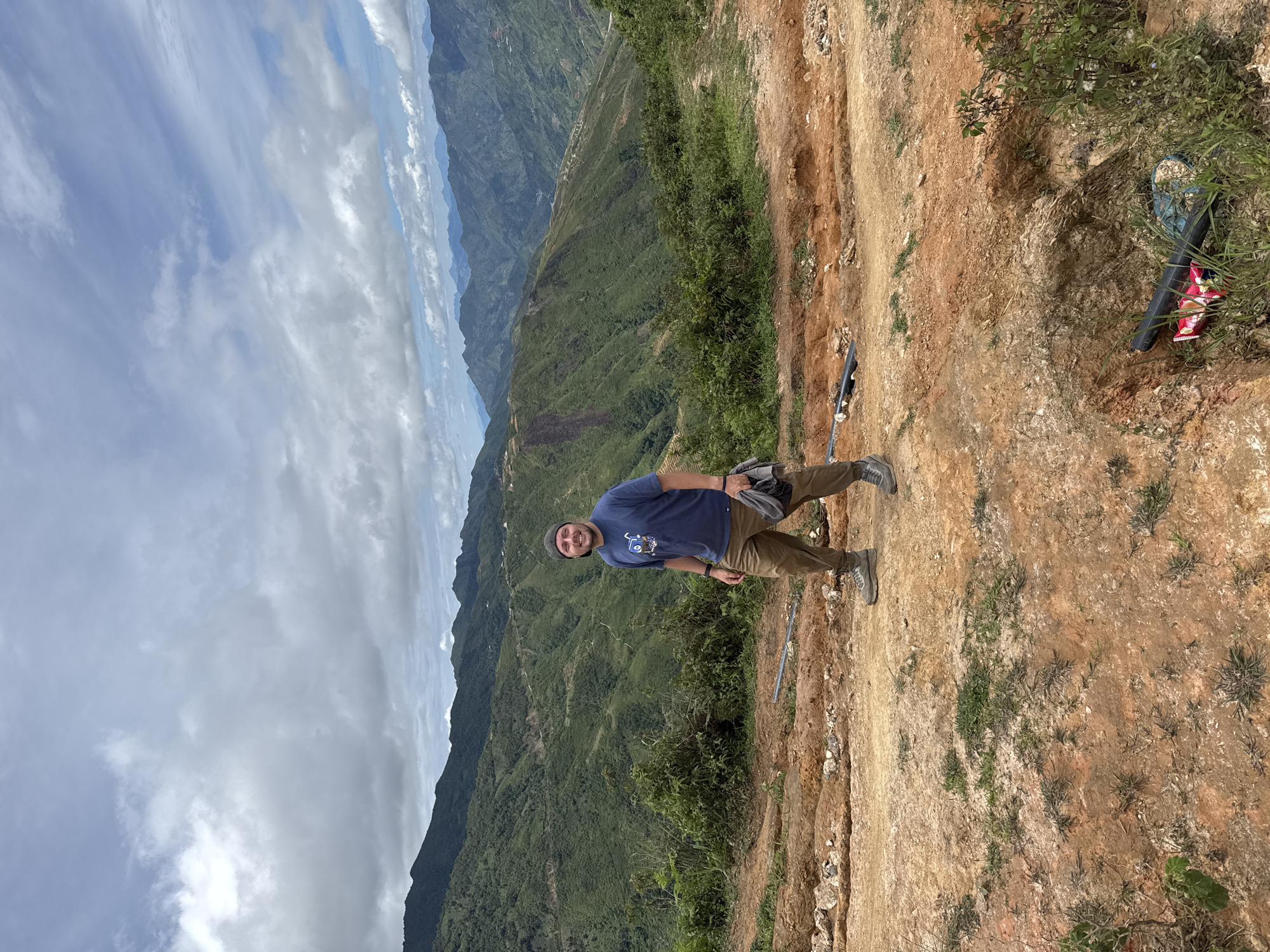 A man stands on a dirt path with a mountainous, green landscape in the background under a cloudy sky. He is wearing a blue t-shirt with a graphic print, tan pants, and a black baseball cap. His left hand is holding a gray jacket or bundle of clothes. The path appears to be under construction or a rough trail, with exposed earth and some scattered rocks and debris, including some black piping. The mountains are covered in dense vegetation, with visible roads winding up their slopes. The overall setting suggests a rural, possibly remote, location.