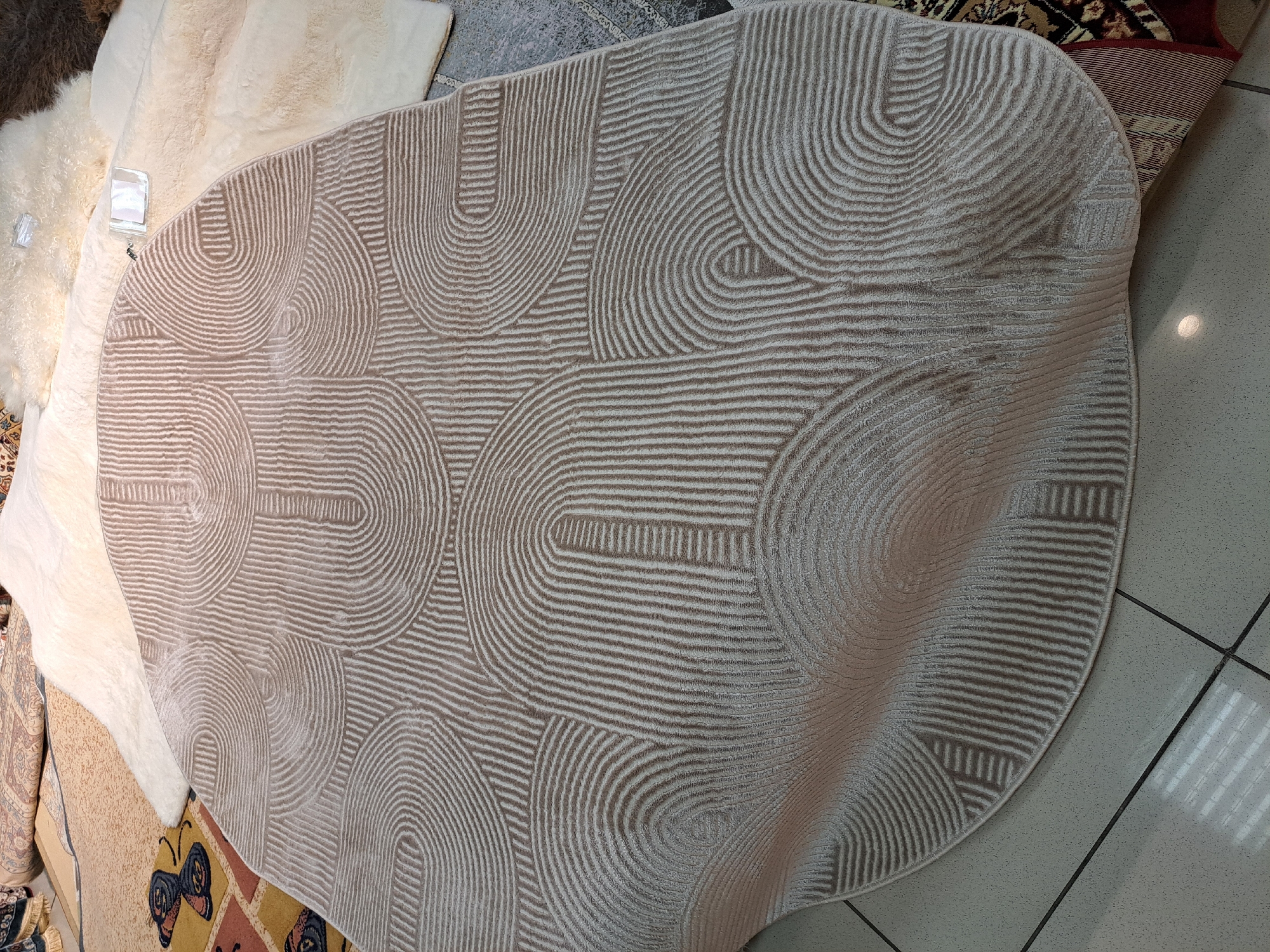 The image shows a beige rug with a geometric pattern of curved lines and rectangular shapes. The rug is partially unrolled and lies on a tiled floor. Several other rugs are visible in the background, including a fluffy white rug and a rug with a butterfly pattern. The scene appears to be a rug store or display area. The location is identified as Barnaul, Russia. There are no people visible in the image.