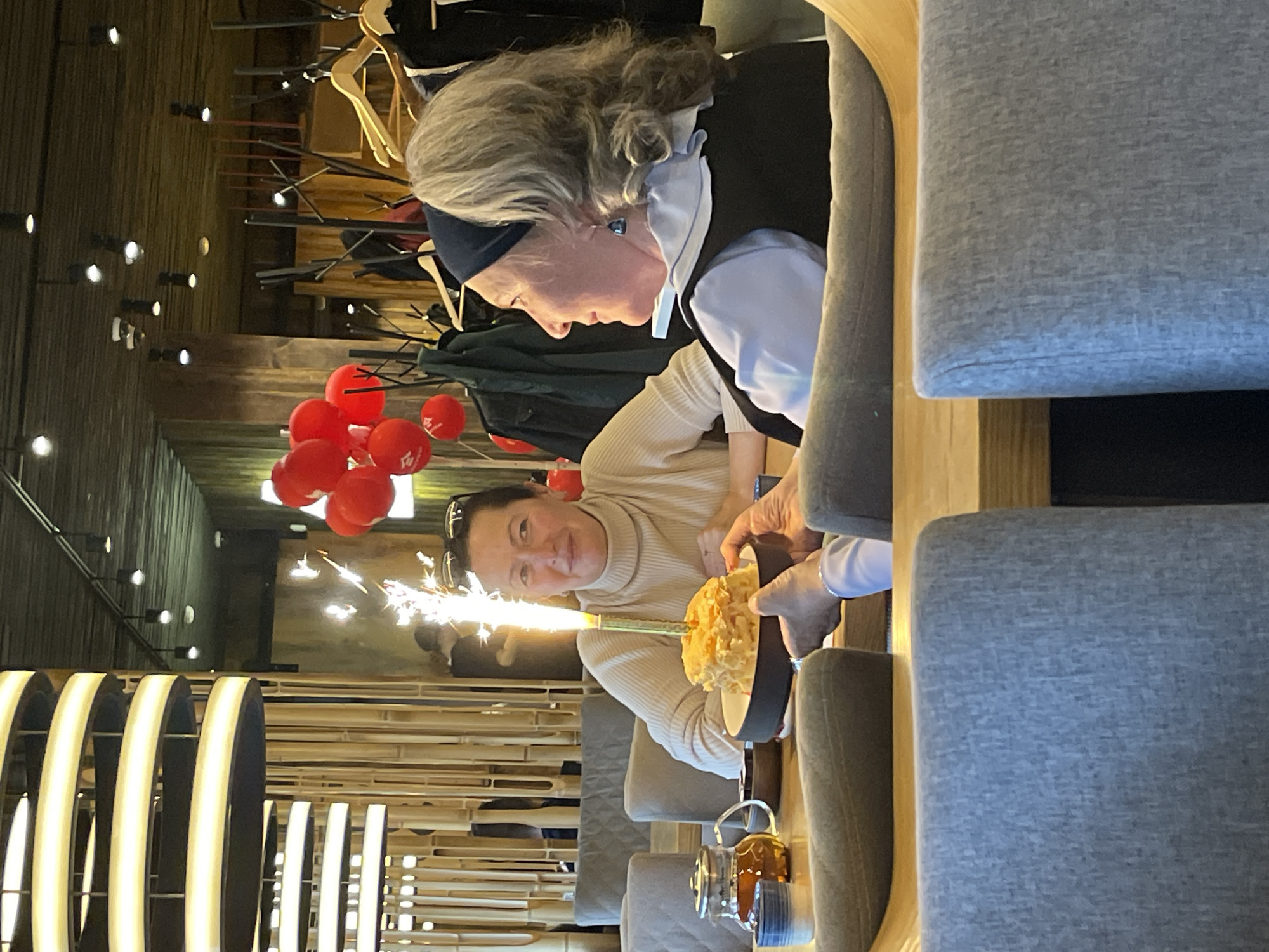 In a restaurant with bamboo-paneled walls and modern lighting, two women are seated at a wooden table. The woman on the left, with dark hair and wearing a cream-colored turtleneck, smiles as she holds a small cake with a lit sparkler. The woman on the right, with gray hair and wearing a black vest over a blue shirt, looks at the cake. Red balloons are visible in the background, suggesting a celebration. A teapot and cups are on the table. The scene appears to be in Moscow, Russia.