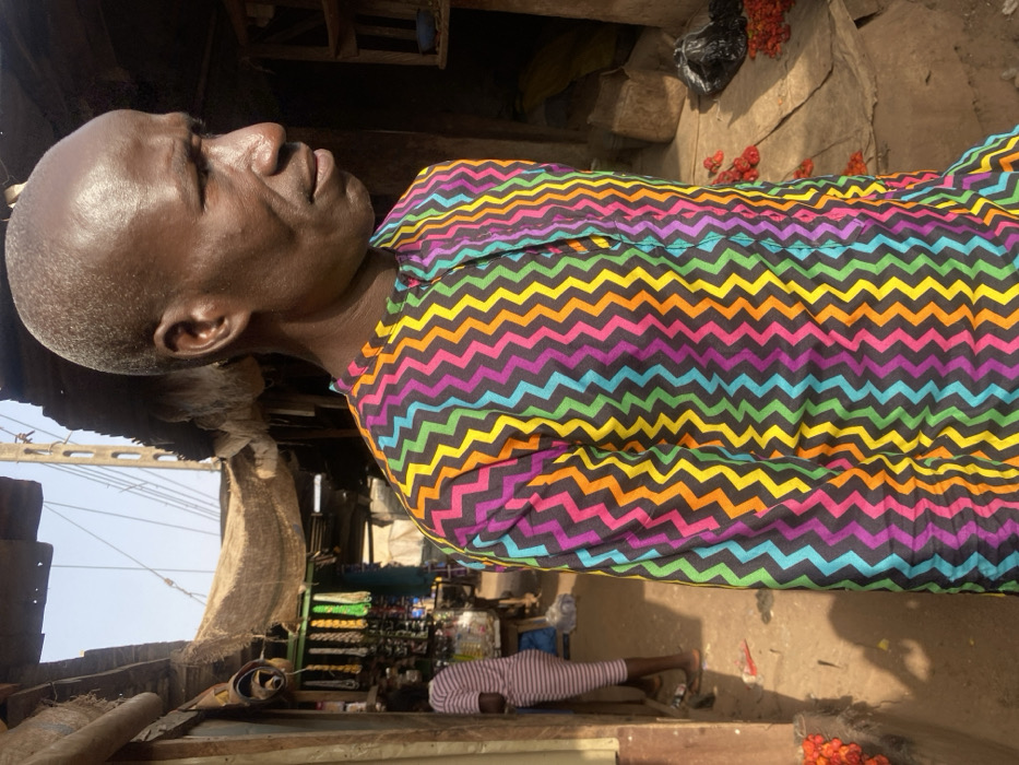 An elder African man with a shaved head is photographed outdoors, wearing a shirt with a chevron pattern of black, orange, yellow, green, blue, and purple. He's positioned at an angle, looking to the left. The background shows a market scene with various goods displayed, including shoes. Another person in a striped outfit lies on the ground. Additionally, there are red fruits or vegetables scattered around on the ground. The scene suggests a bustling, everyday life in an African marketplace.