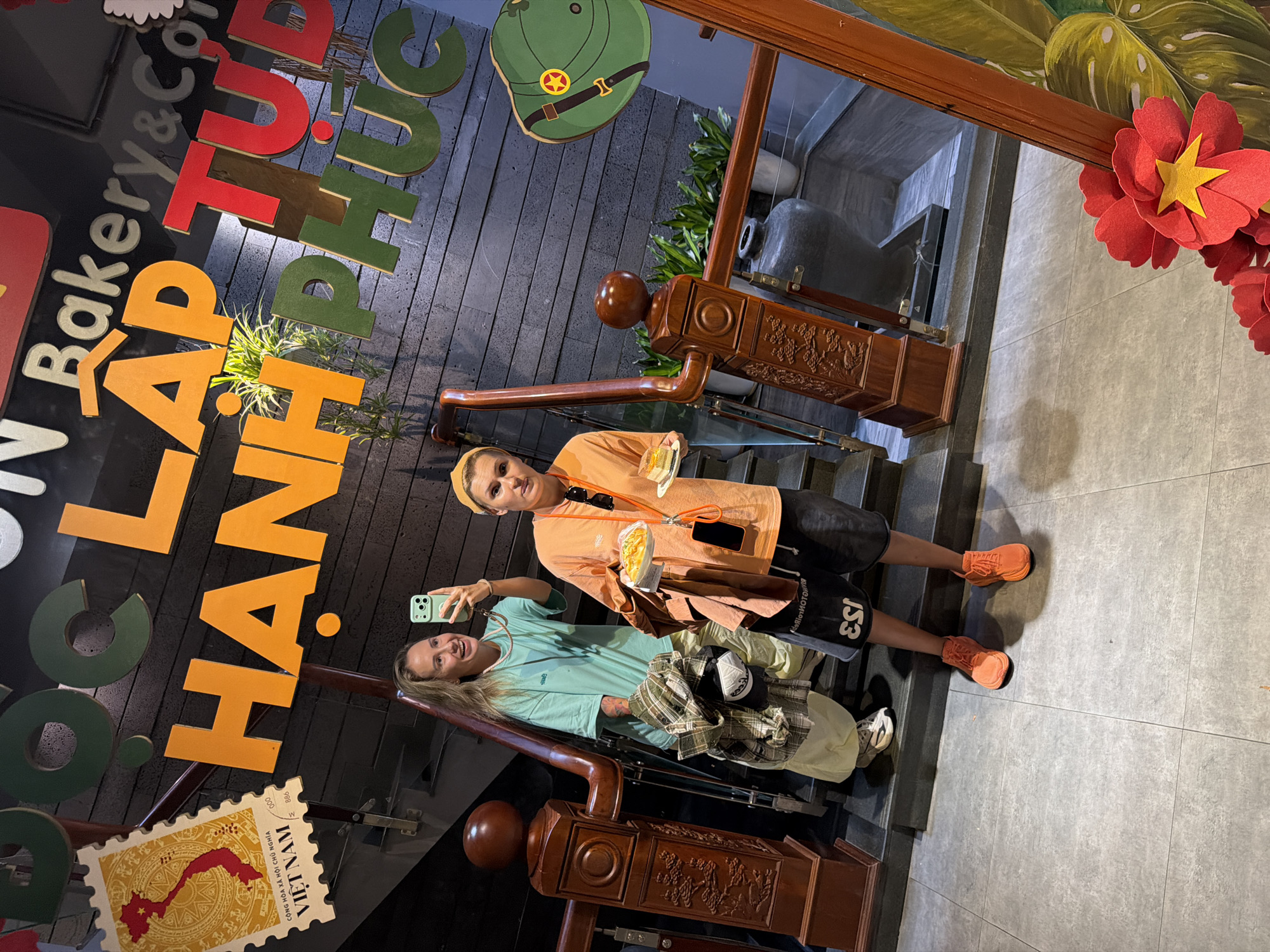 Two individuals are positioned on a wooden staircase with a grey-tiled floor. The person on the left, wearing a teal t-shirt, is holding a smartphone and taking a selfie. They are carrying a plaid shirt and a cap. The person on the right, in an orange t-shirt, holds a plate with two pieces of cake. They are wearing black shorts and orange sneakers. A large wooden railing with carved details runs along the staircase. Above them, large, colorful letters spell out "C LAP TU HANH PHUC" on a dark, textured wall. A stamp-like graphic of Vietnam is visible on the left side of the wall. Further up the stairs, potted plants and a large dark vase are present. The scene appears to be a well-decorated indoor space, potentially a cafe or restaurant, in South Quy Nhon Ward, Vietnam, given the visual cues.
